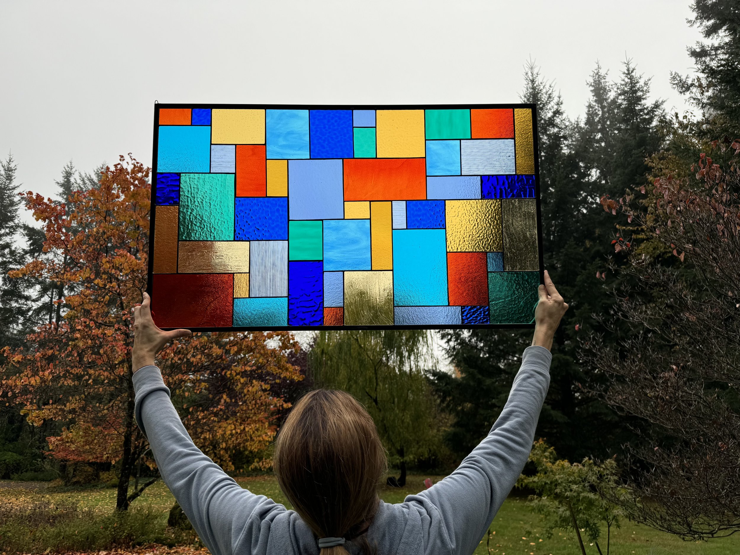 Person holding a colorful stained glass window outdoors with trees and autumn foliage in the background.