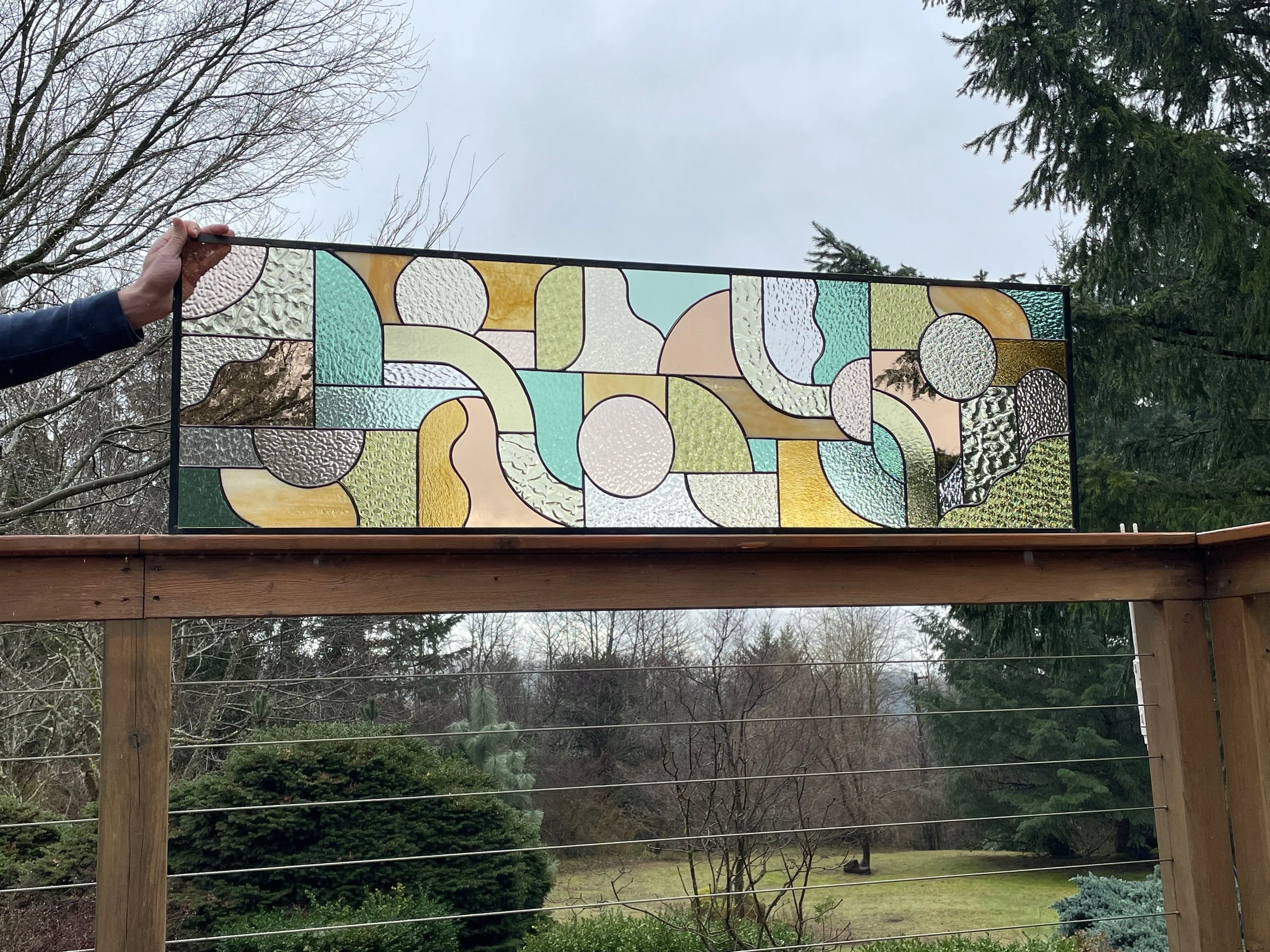 A person holding a stained glass panel with abstract geometric shapes and textured glass in shades of green, yellow, cream, and white on a wooden railing outdoors, with trees and an overcast sky in the background.