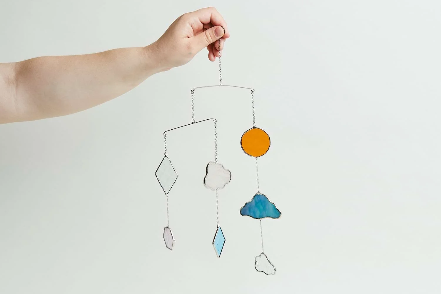 Hand holding a hanging mobile with geometric and cloud-shaped stained glass elements against a plain background.