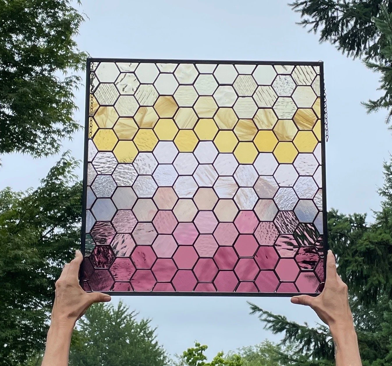 Person holding a square stained glass window with hexagon shapes, featuring yellow, pink, purple, and clear glass sections, outdoors with trees and a cloudy sky in the background.
