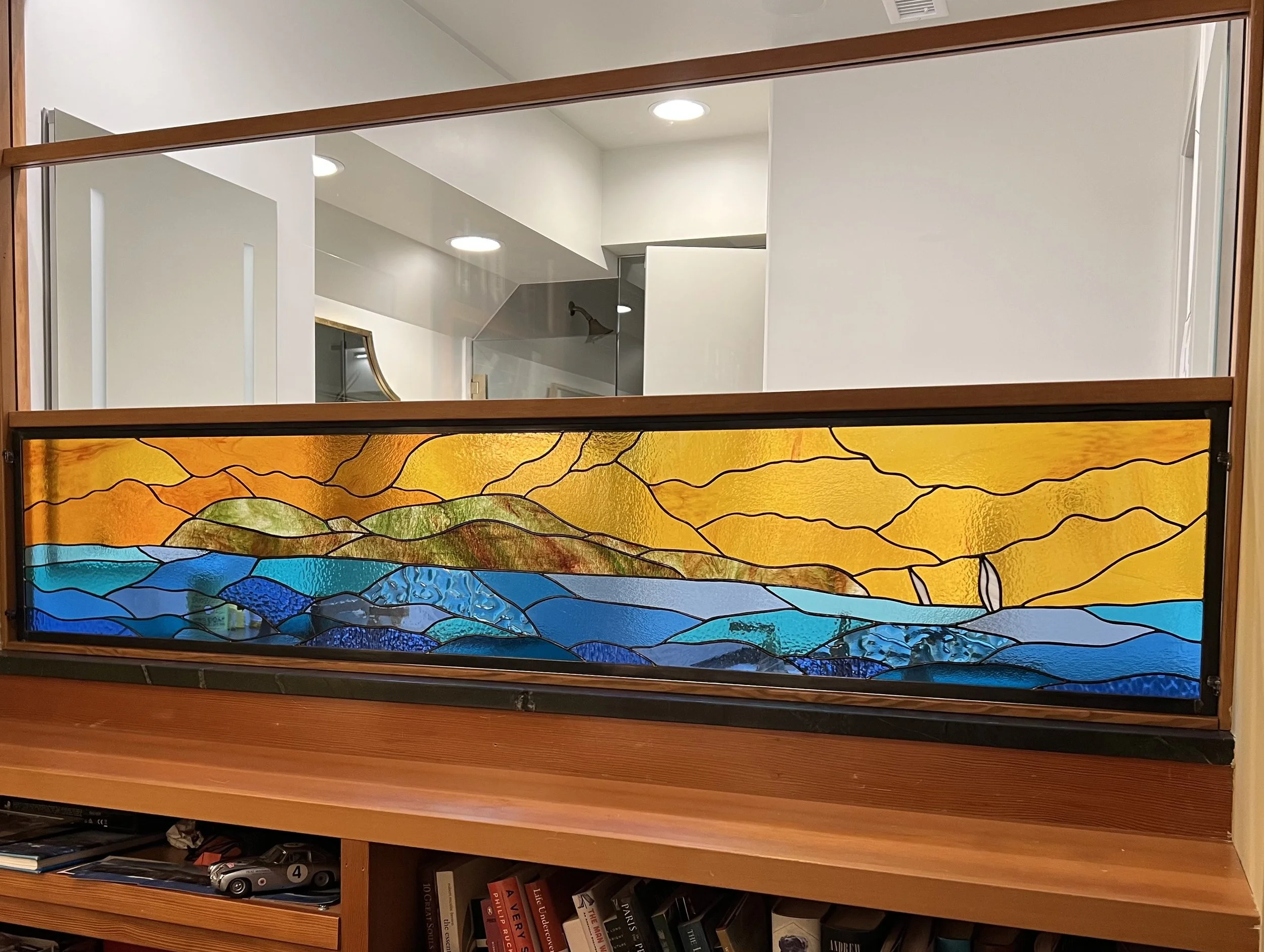 A stained glass landscape artwork depicting mountains, water, and a sky with orange and yellow hues, placed on a wooden cabinet with books and decorative items underneath.