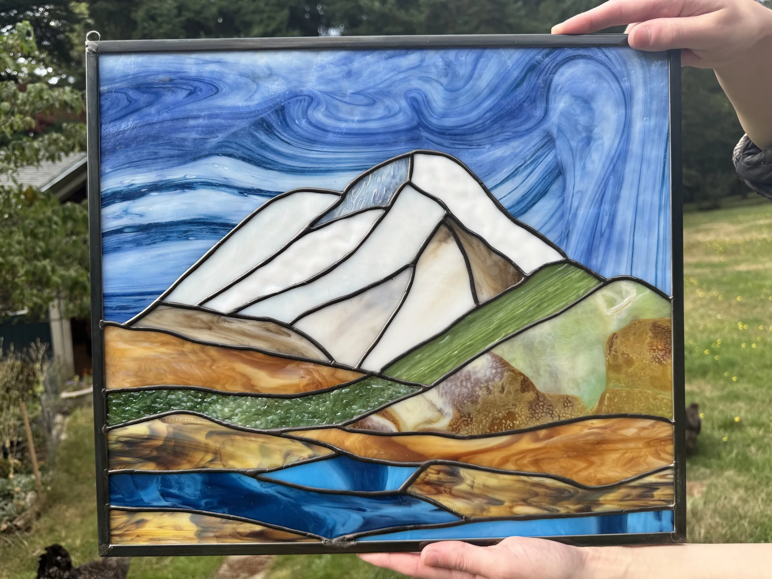 Stained glass window depicting a mountain landscape with snow-capped peaks, clouds, green hills, and a blue river or lake in the foreground.