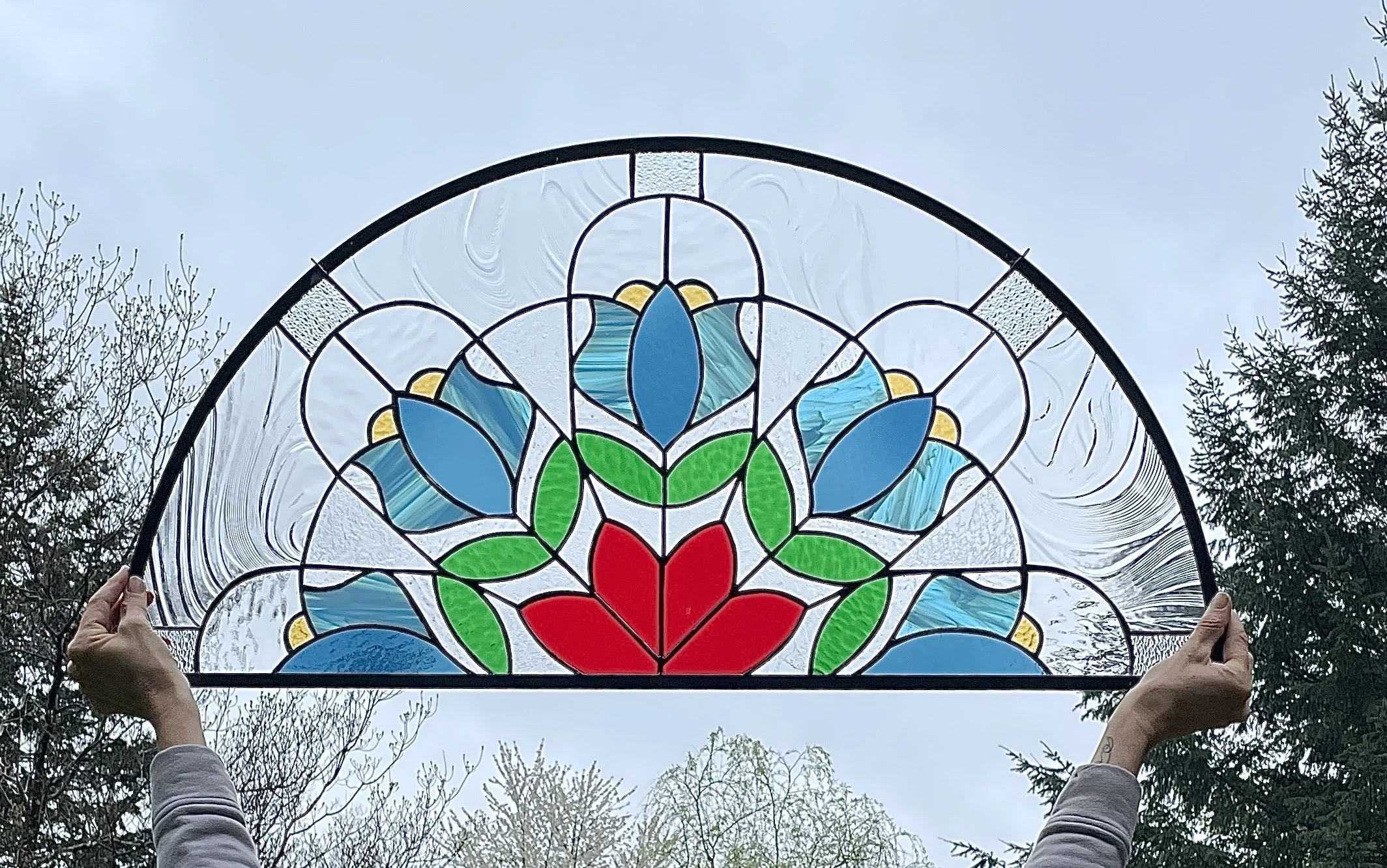 Person holding up a stained glass window with blue, red, green, and yellow floral patterns against an outdoor sky and trees.