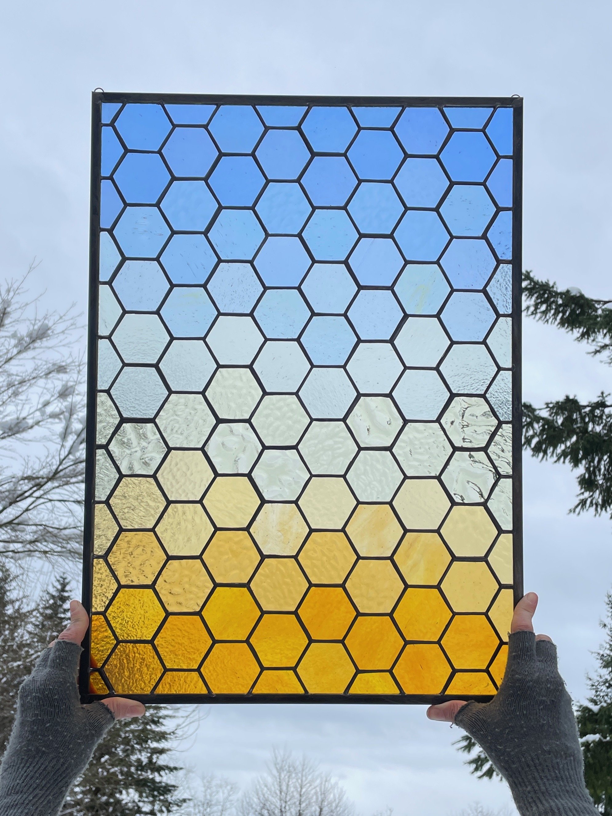 Person holding a stained glass panel with hexagonal pieces, transitioning from yellow at the bottom to blue at the top, against a cloudy sky and trees.