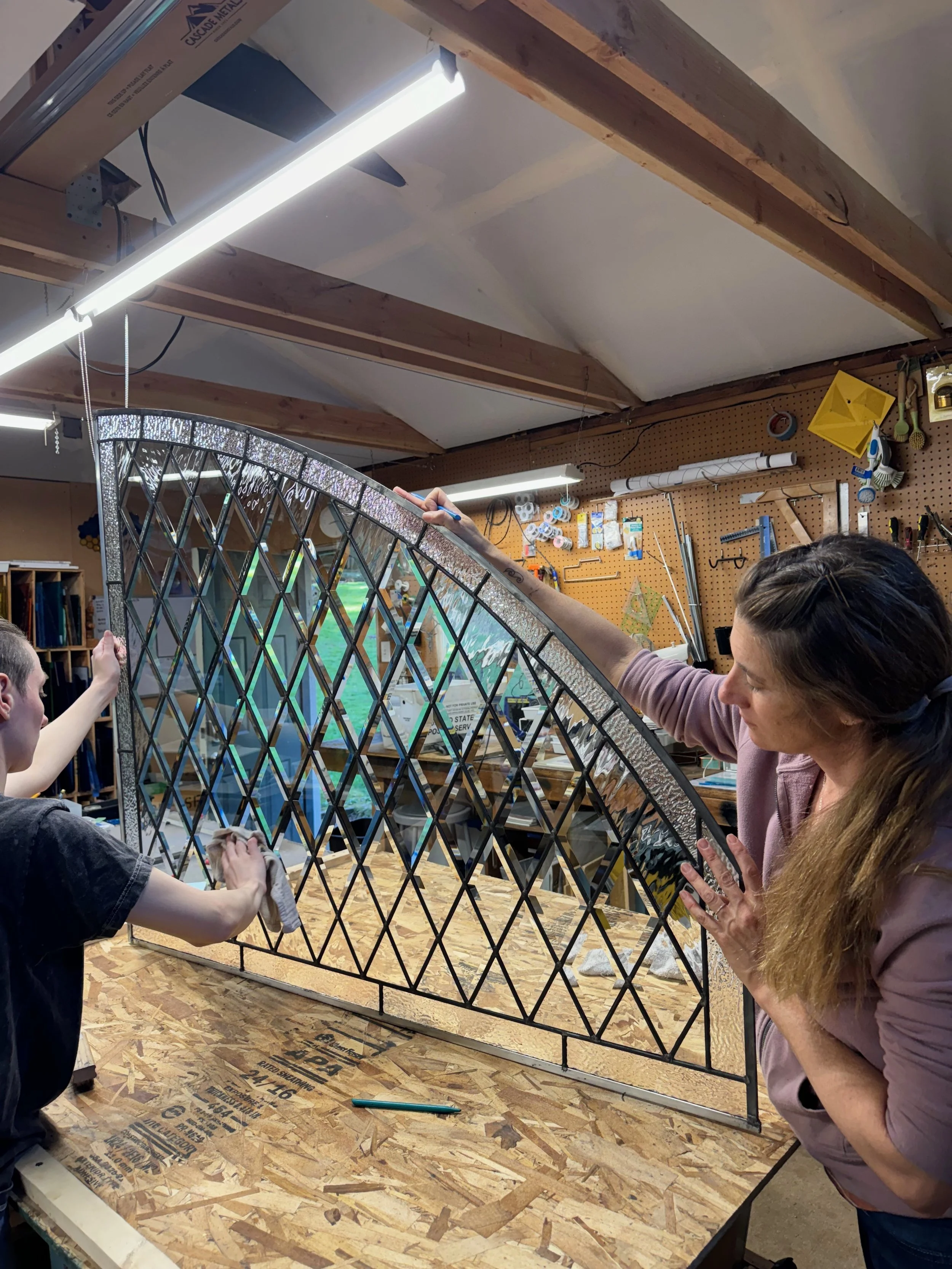 Two women working together on a stained glass window in a woodworking workshop, with tools and materials hanging on the wall behind them.