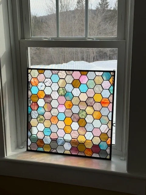 Colorful stained glass panel with hexagonal patterns placed on a windowsill, seen from inside a room with snow visible outside.