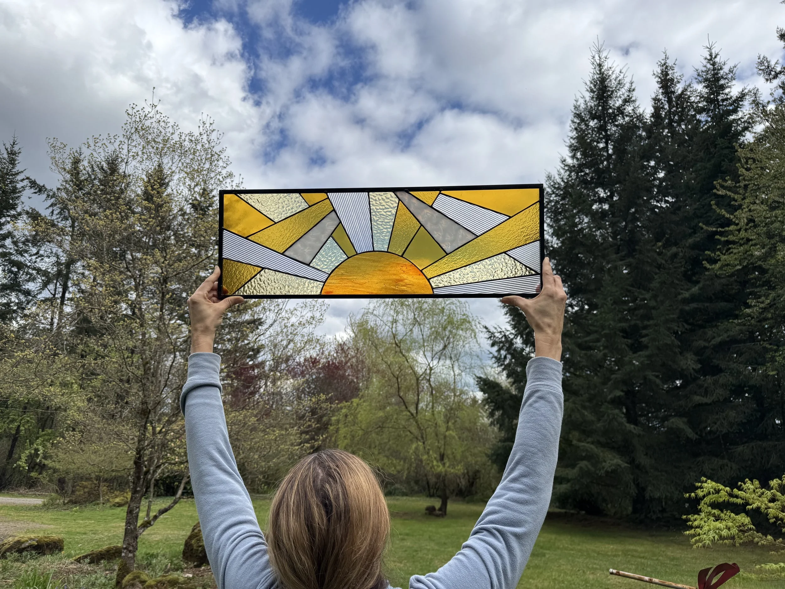 A woman in a gray long sleeve shirt holding a stained glass panel depicting a stylized sunrise with rays spreading across the sky, outdoors in a park with trees and a cloudy sky in the background.