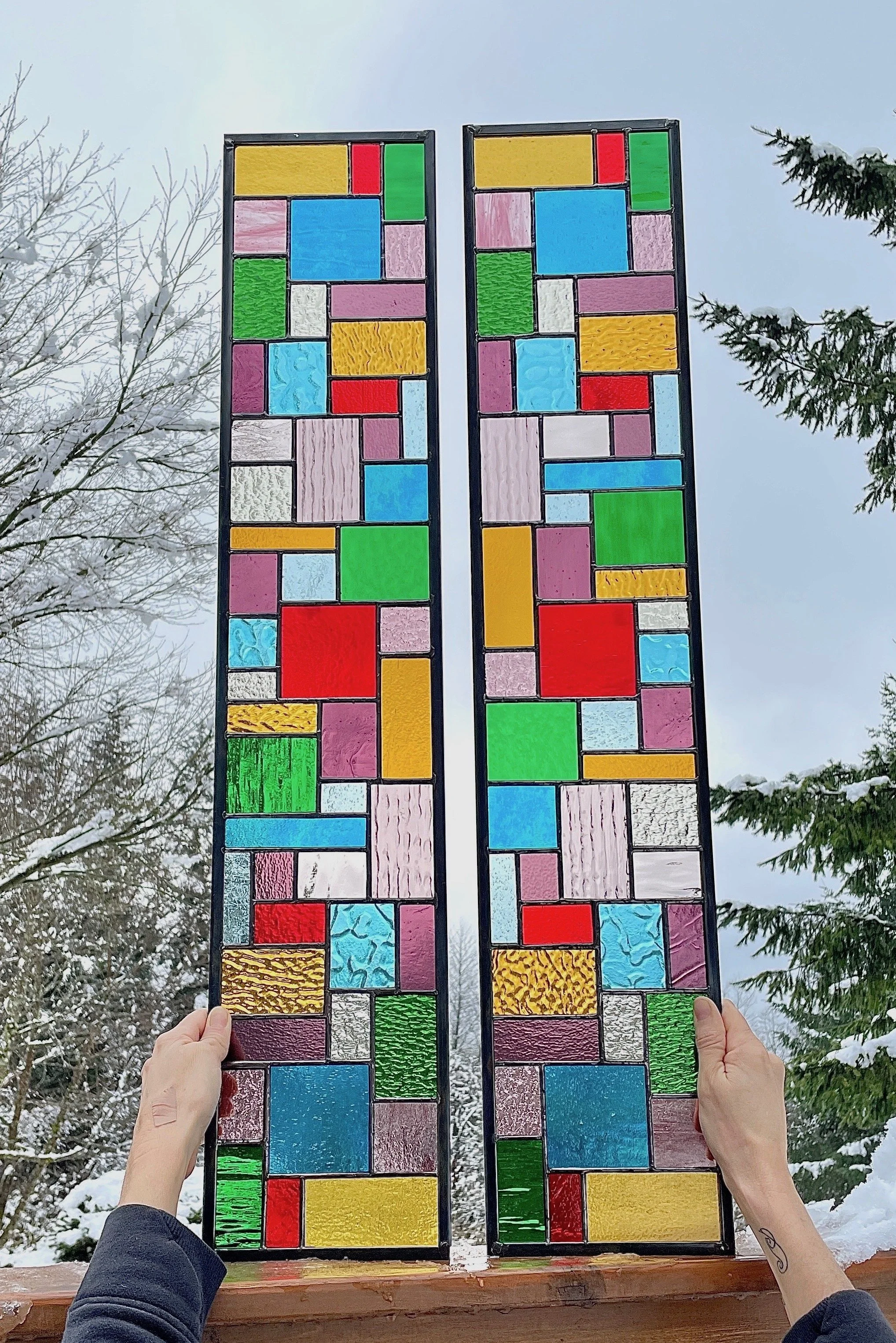 Two stained glass panels with colorful geometric patterns being held outdoors over a snowy landscape.