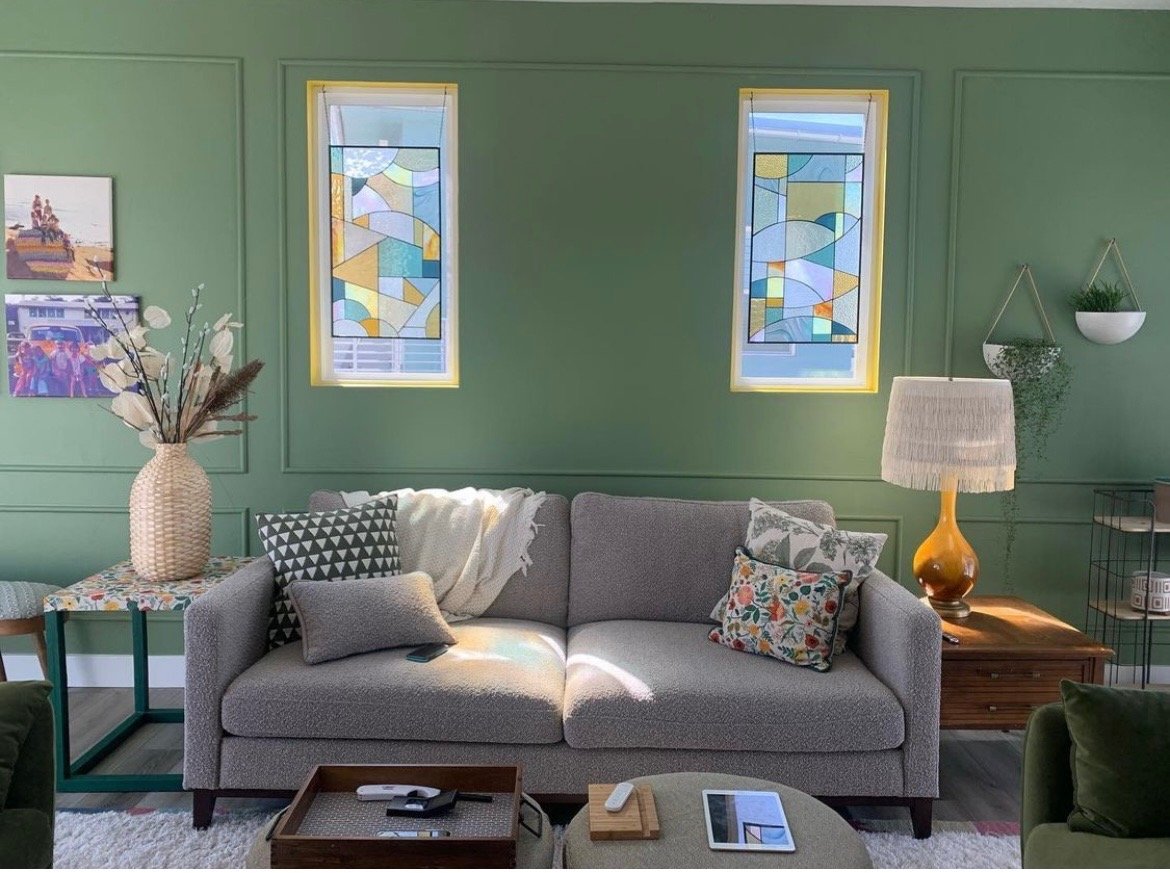 Living room with green wall, three stained glass windows, gray sofa with patterned pillows, wooden side table with lamp, decorative items, and artwork on the wall.