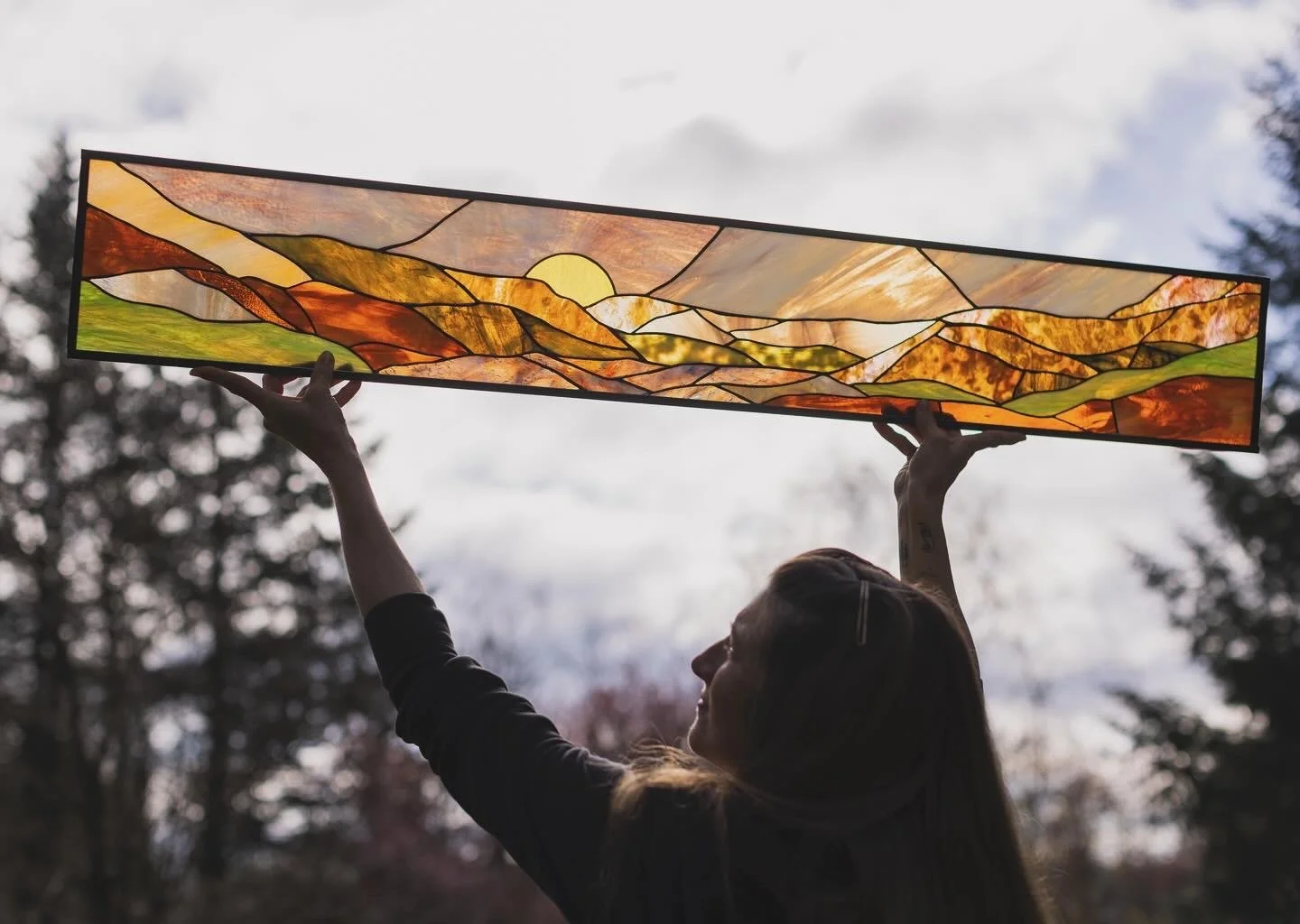 A person holding a large rectangular stained glass panel depicting a landscape with mountains, a setting sun, and a sky with clouds, outdoors during dusk or dawn.