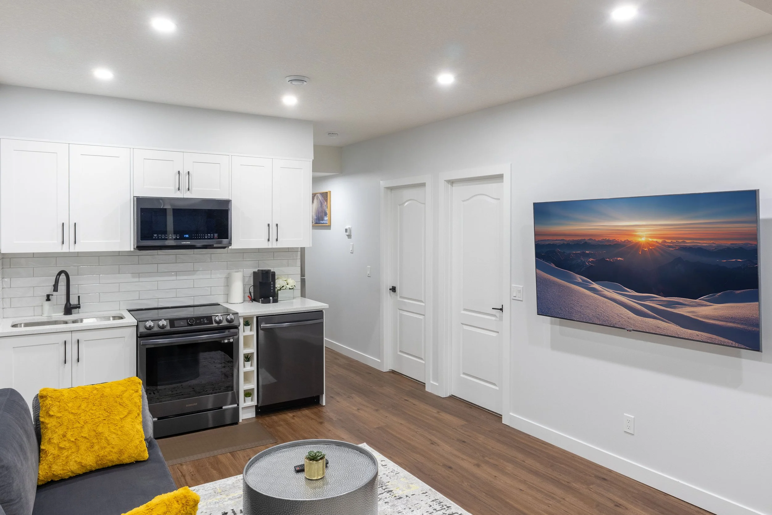 Modern basement with gray velvet sofa, yellow fuzzy pillows, hammered metal coffee table, white kitchen cabinets, black appliances, and wood flooring.
