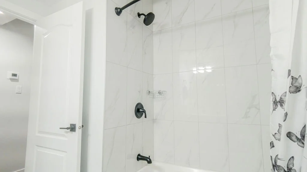 bathroom with glossy white marble-veined tiles, matte black shower fixtures, white tub, and a butterfly-print curtain in bright indoor light.