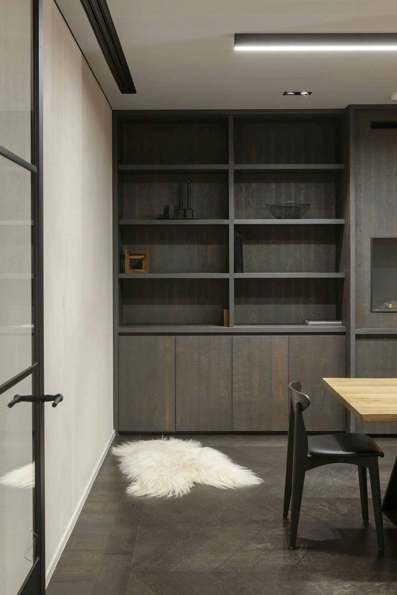 Modern interior room with dark wooden built-in shelves, a white fluffy rug on dark wood flooring, part of a table and a black chair visible.