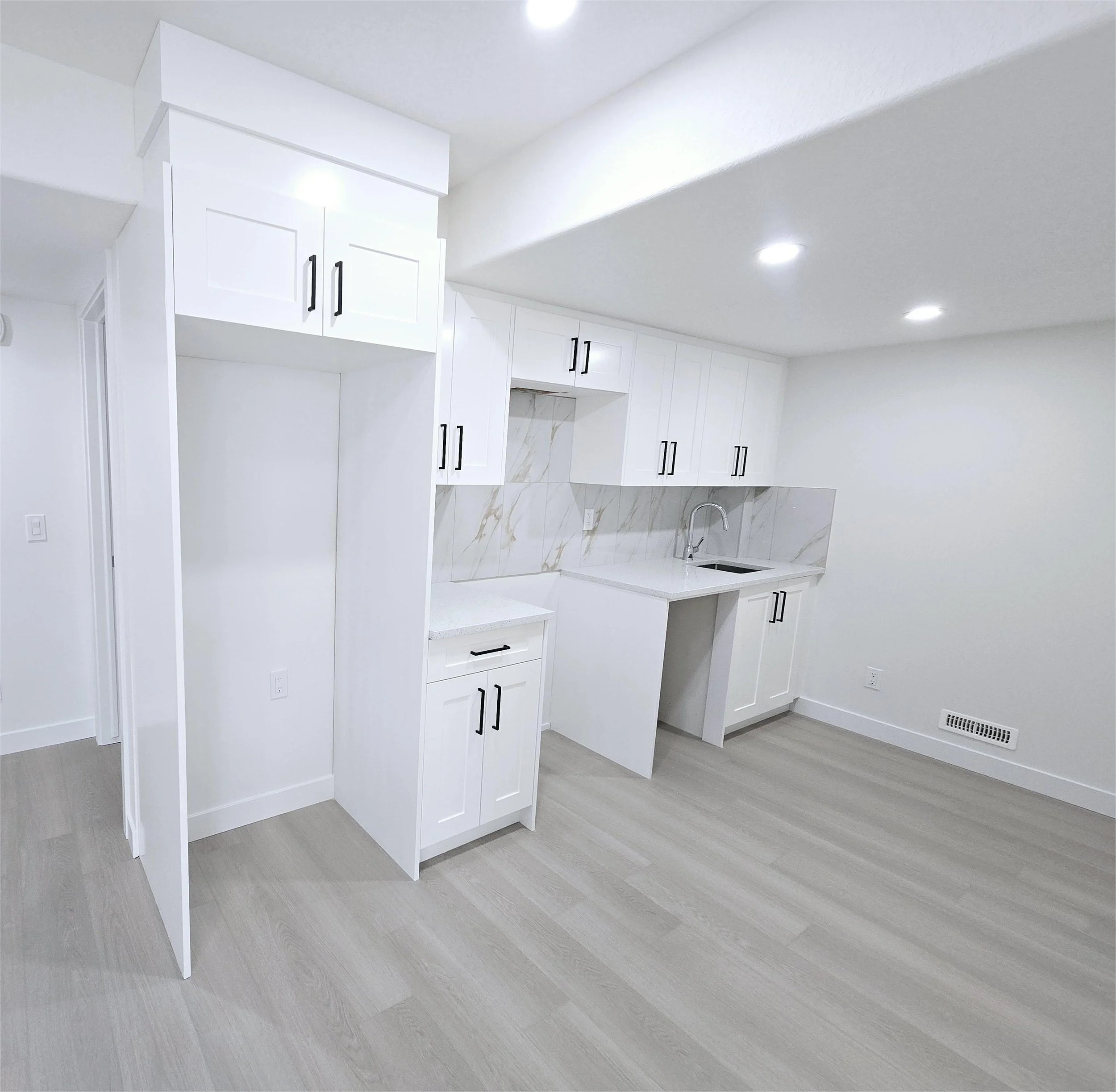 Minimalist white kitchenette with shaker cabinets, black hardware, marble backsplash, grey plank floors, and bright recessed lighting.