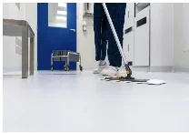 Medical worker in blue scrubs using a flat mop on a white sterile hospital floor, near a vibrant blue door, metal cart, and clinical equipment.