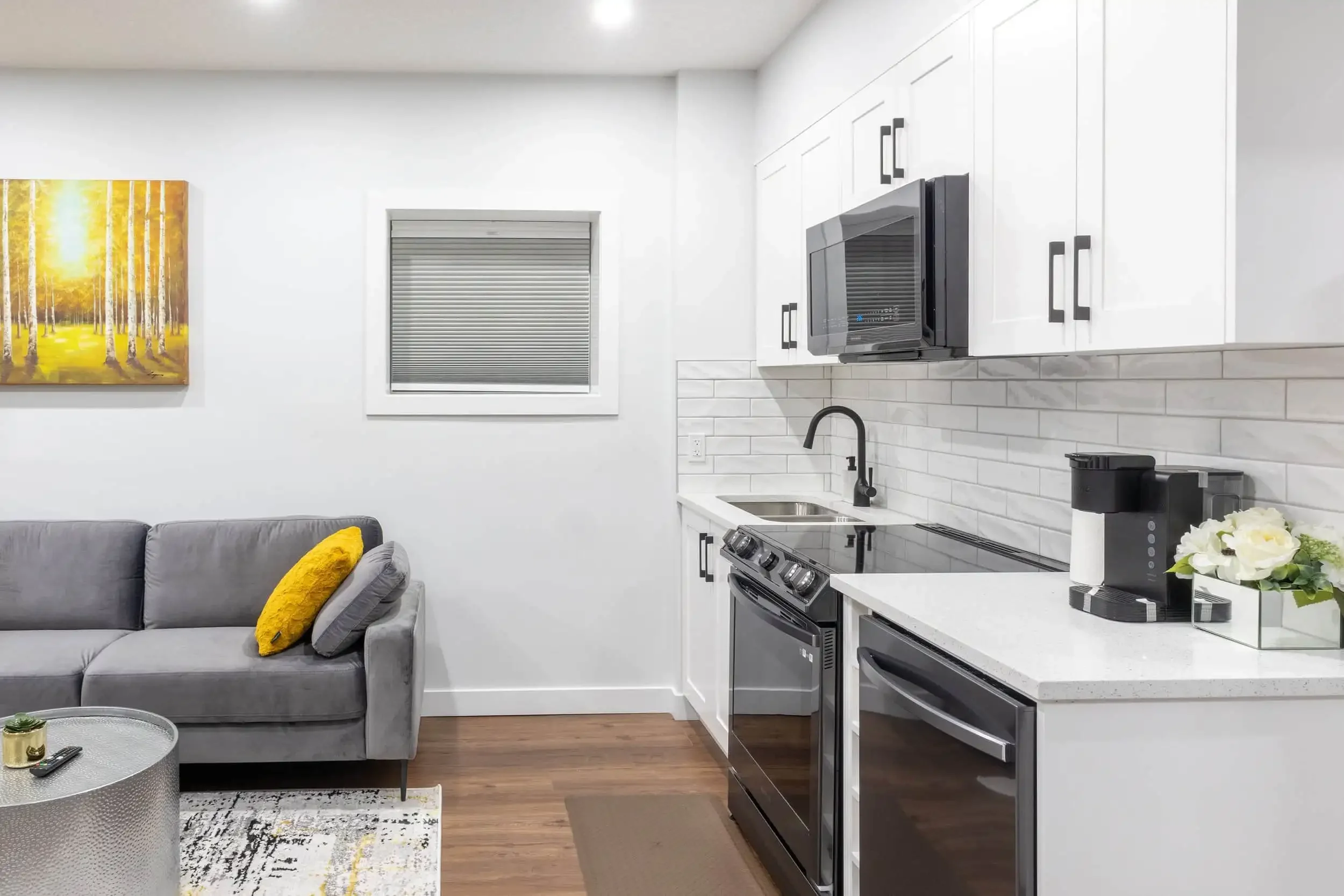 Modern white kitchen with quartz counters, black hardware, subway tile, gray velvet sofa, and yellow forest art in a bright studio apartment.