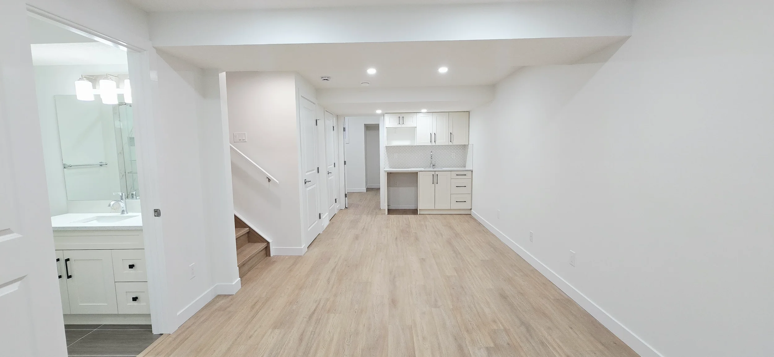 Bright modern basement suite with light oak vinyl plank flooring, white shaker kitchenette, recessed pot lights, and crisp white walls and doors.