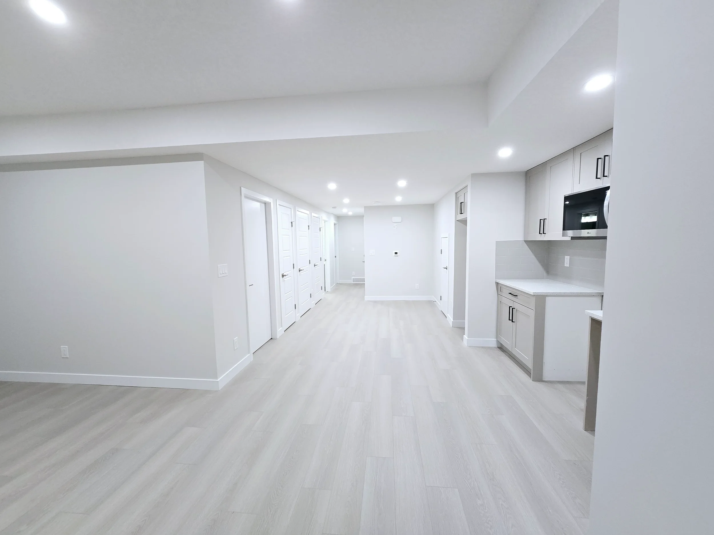 Bright modern basement featuring light gray wood-look flooring, white walls, recessed lights, and a compact kitchenette with gray shaker cabinets.