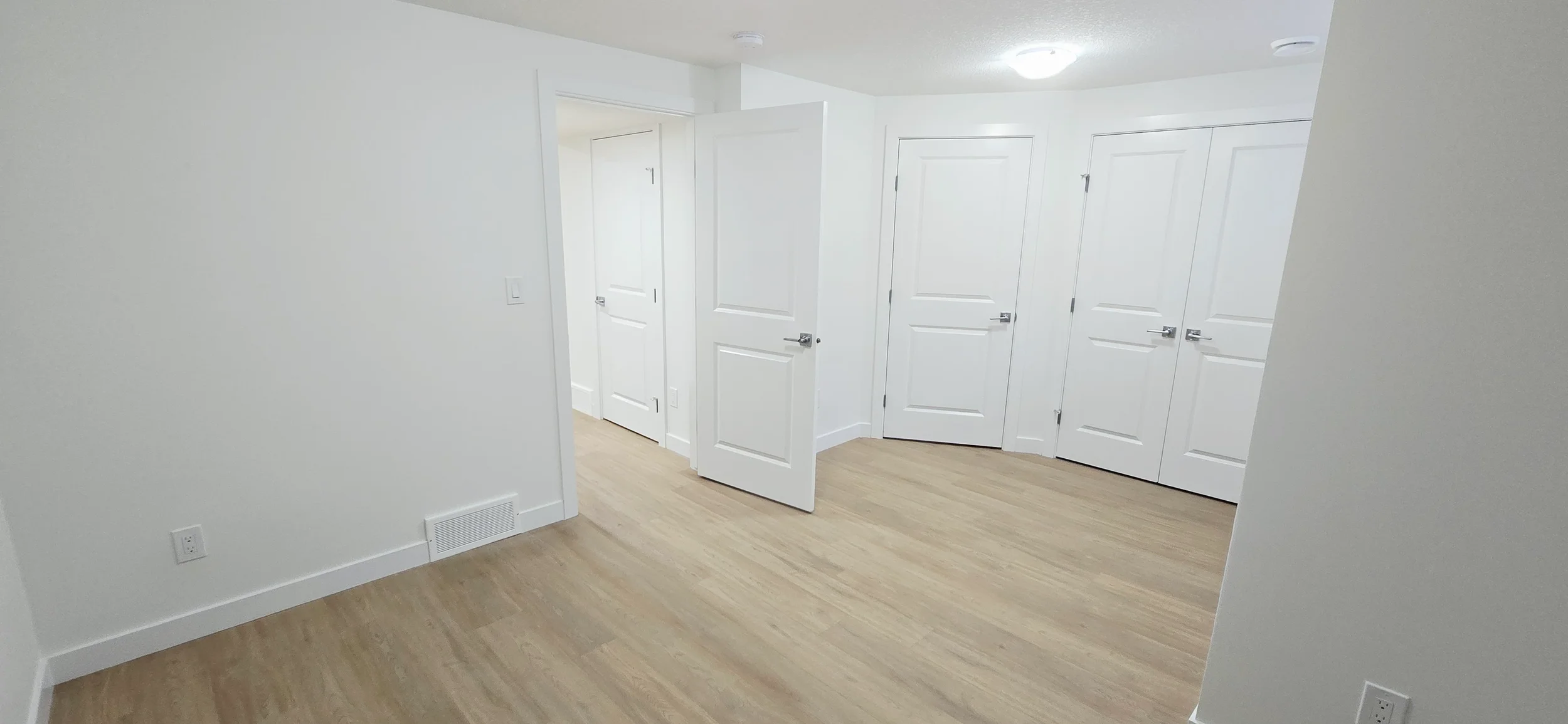 Modern empty room with white walls, light oak wood-look floors, multiple white doors, and bright ceiling light. Clean minimalist home interior.
