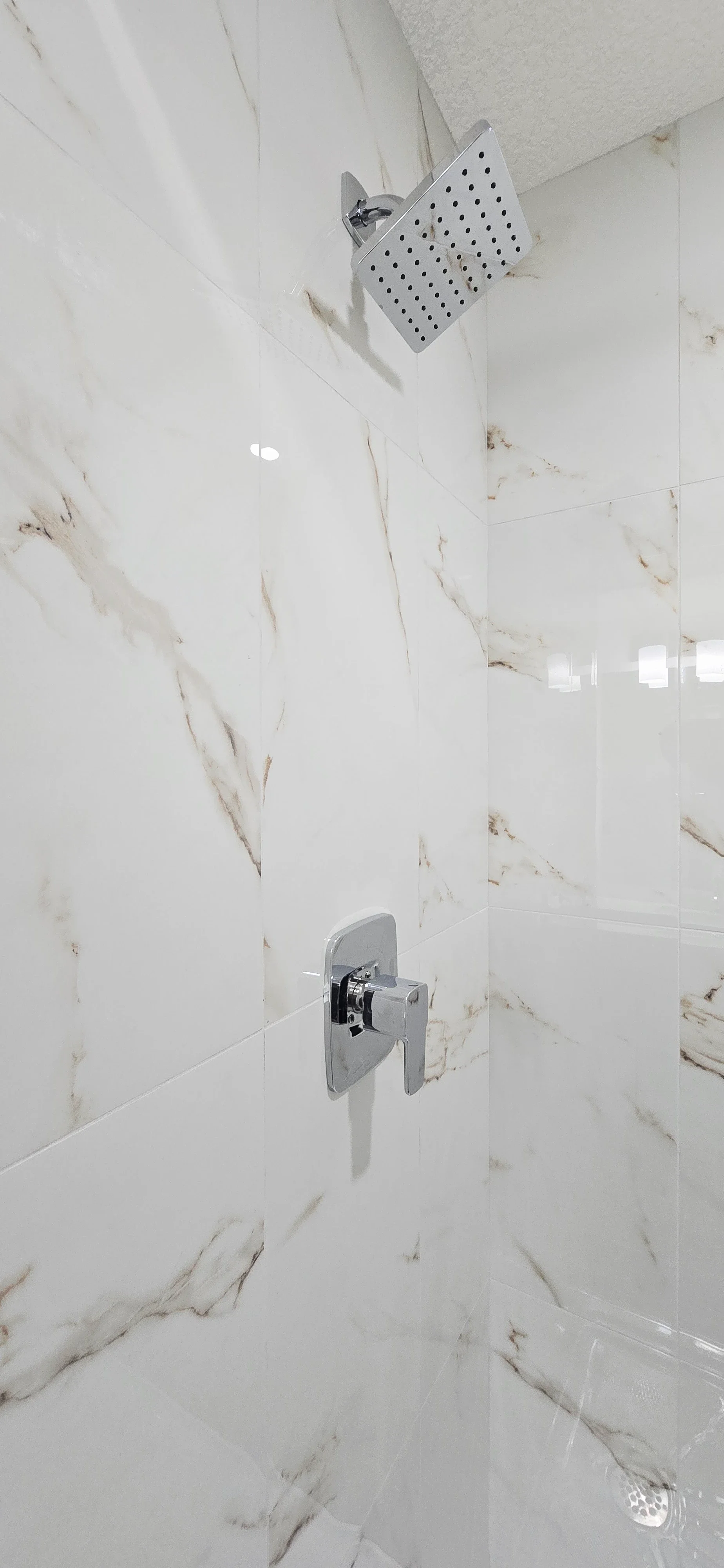 Bright modern shower featuring polished chrome rainfall head and handle set against glossy white marble-patterned tiles with gold veining.