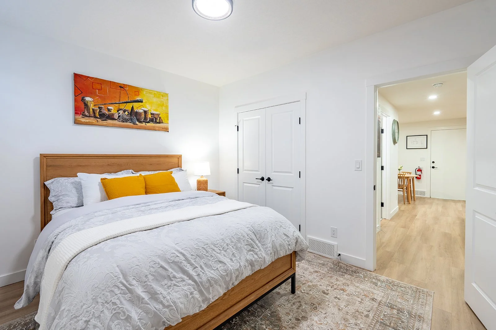 A bedroom with a white bed, yellow pillows, wooden headboard, white walls, a lamp on bedside table, a painting above the bed, beige carpet, and an open door leading to a hallway with a table and chairs.