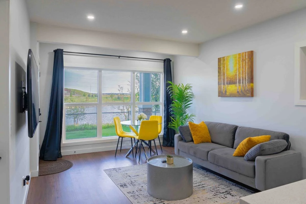 Modern living room with grey velvet sofa, yellow dining chairs, dark blue curtains, lake view window, silver coffee table, and forest wall art.