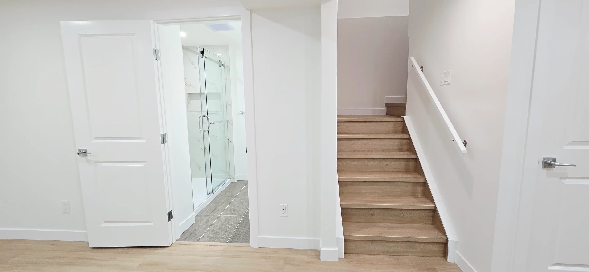 Modern white interior showing an open bathroom door with glass shower, light wood stairs with white handrail, and minimalist neutral decor.