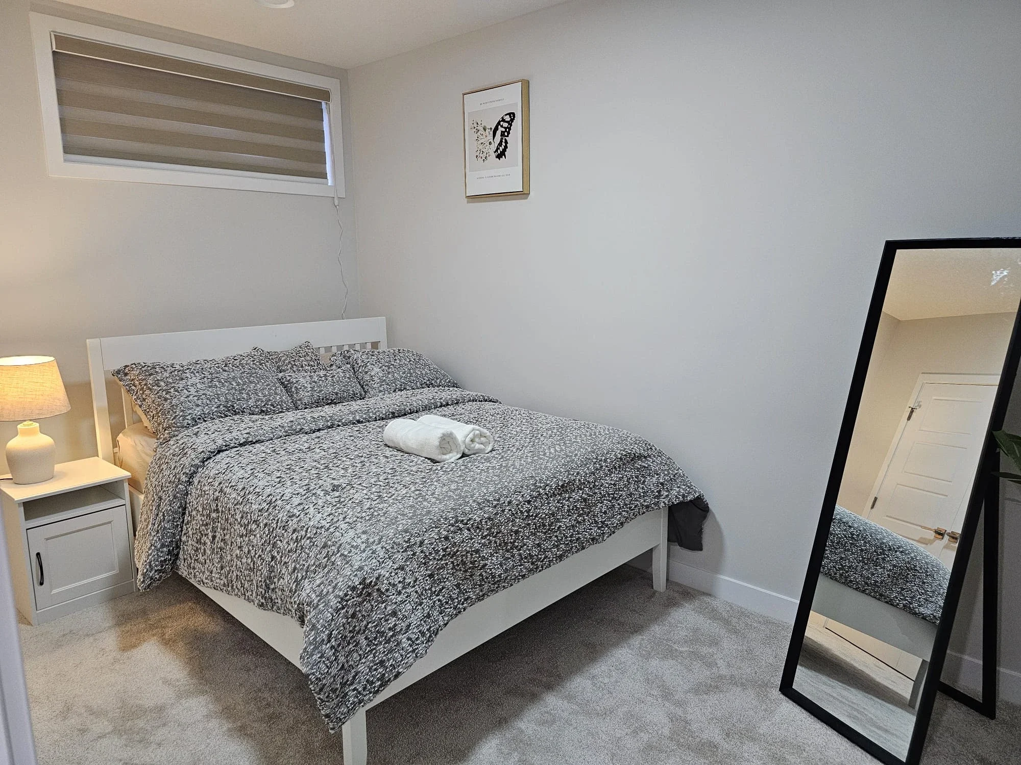 Modern minimalist bedroom with white bed, grey patterned linens, plush carpet, black floor mirror, and warm lighting from a ceramic bedside lamp.