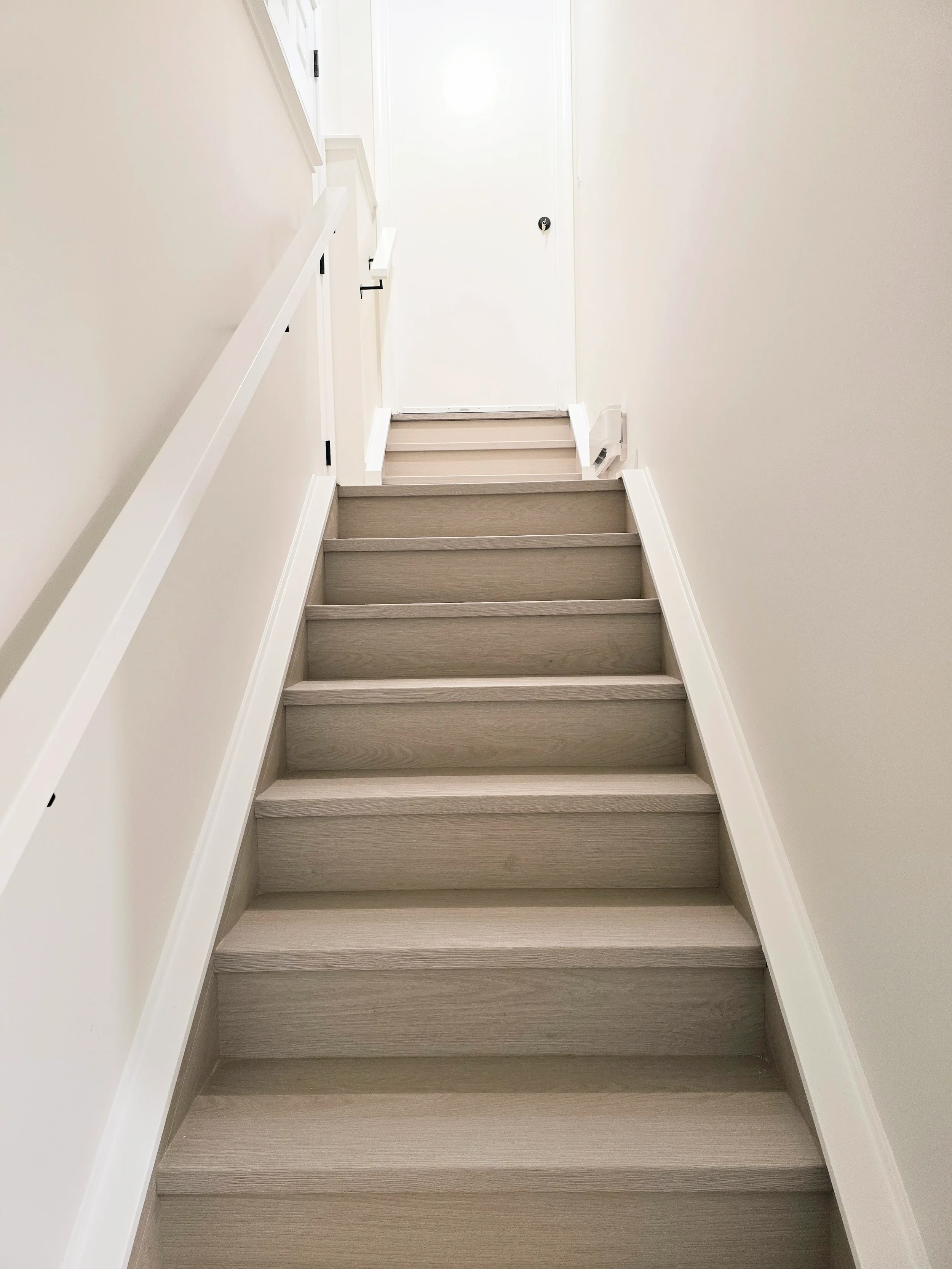 Light-gray wood staircase leads up to a white door in a bright, minimalist hallway with beige walls and white trim and handrail.