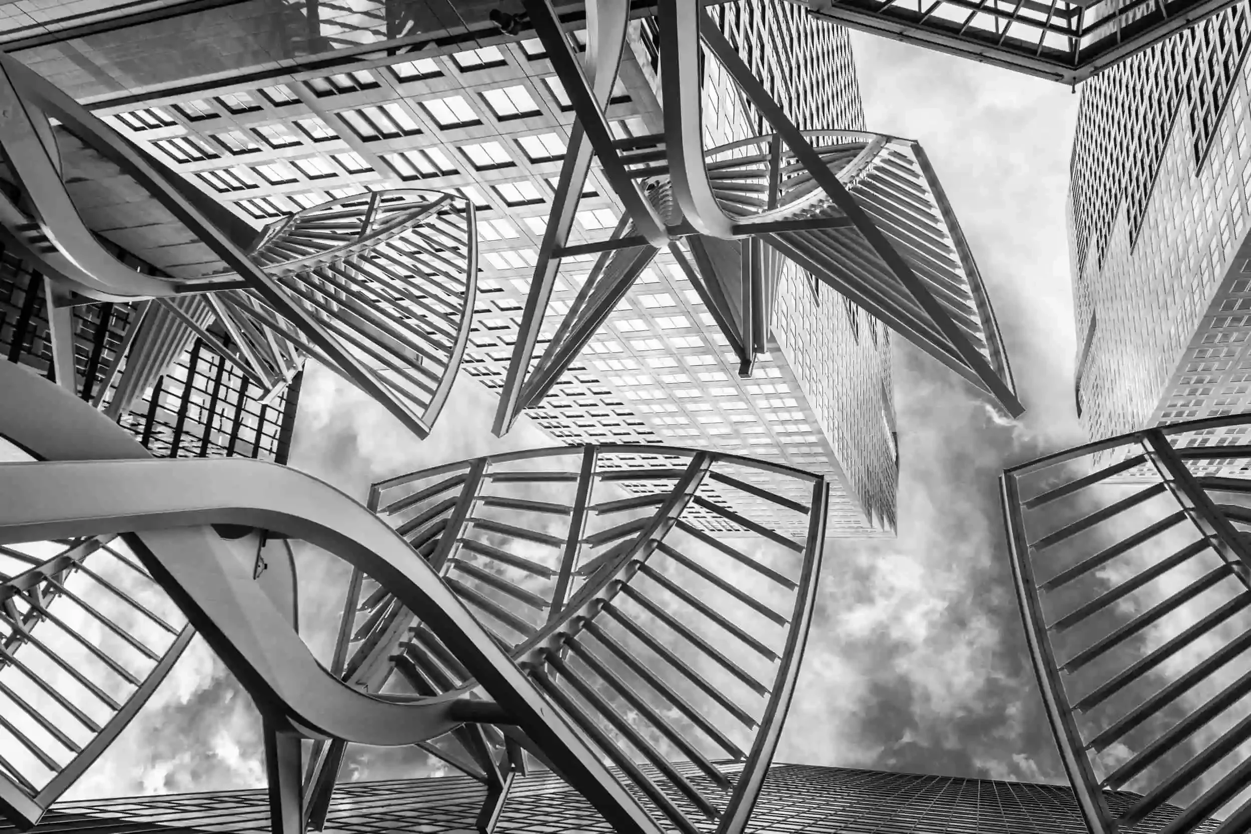 Dramatic black and white low-angle view of curved metallic ribs framing glass skyscrapers and a cloudy sky. Modern urban architectural photography.
