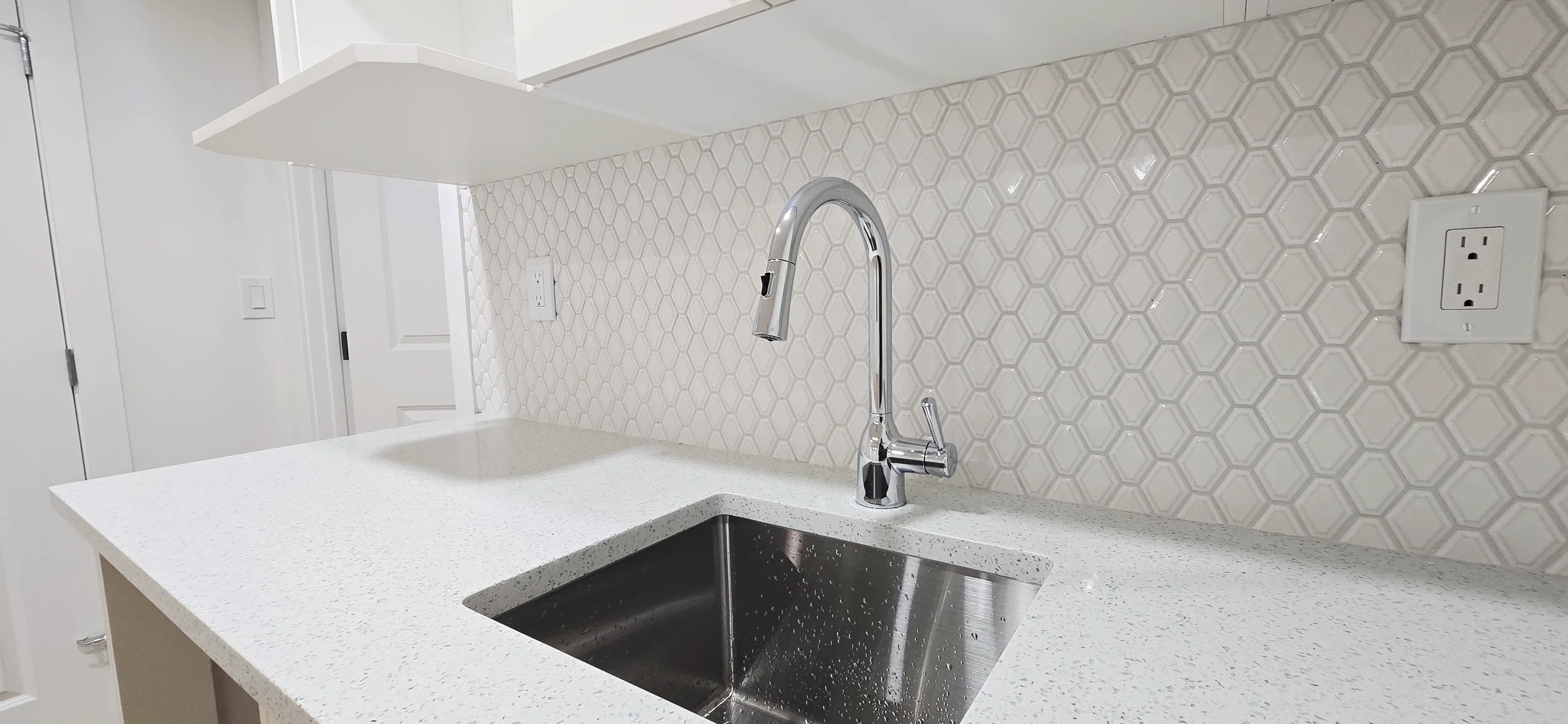 Modern bright kitchen featuring a chrome gooseneck faucet, stainless steel sink, white speckled quartz countertop, and white hexagon tile backsplash.