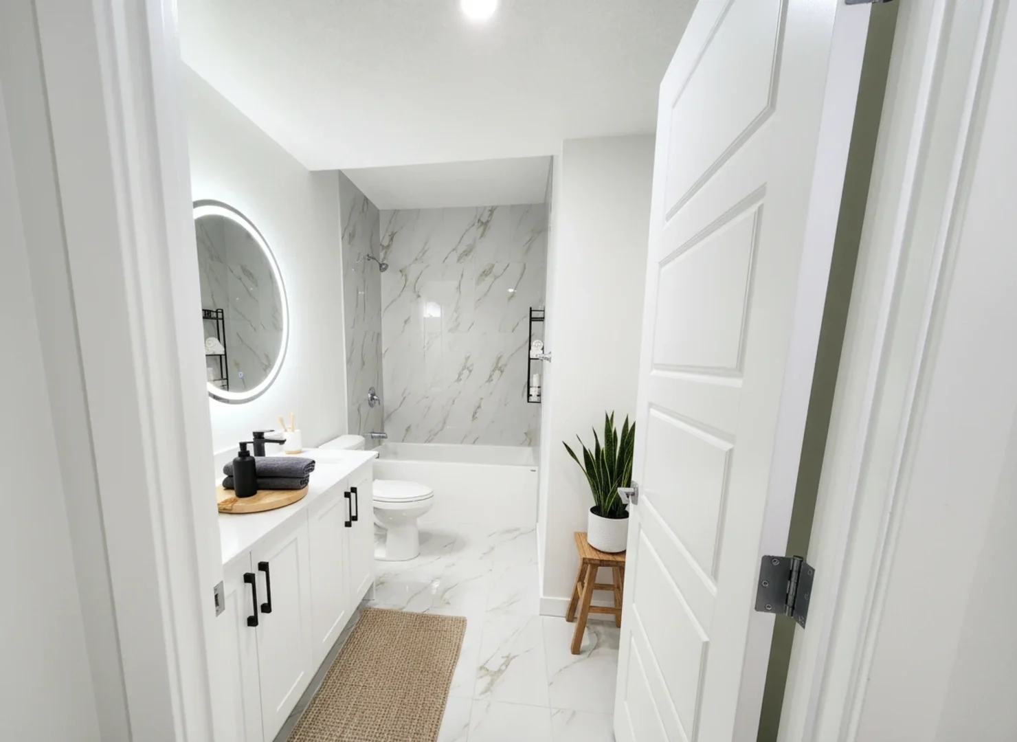Modern, minimalist bathroom with white cabinetry, a round illuminated mirror, marble walls and floors, a potted plant on a small wooden stool, and a bathtub with a shower.