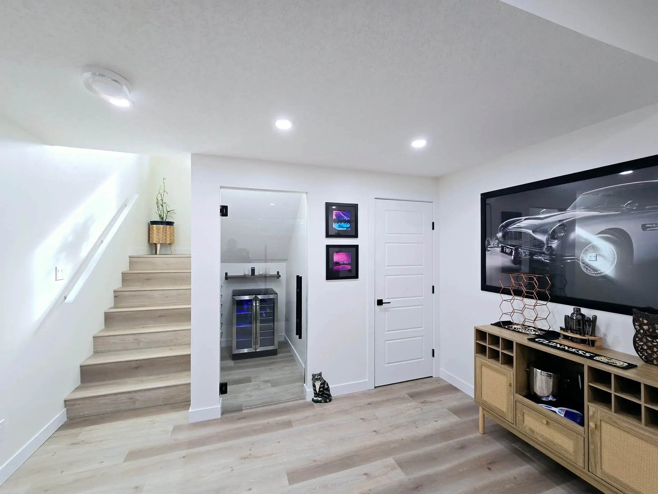 Modern basement with stairs, a small wine fridge, artwork, and a large wall-mounted TV showing a car.