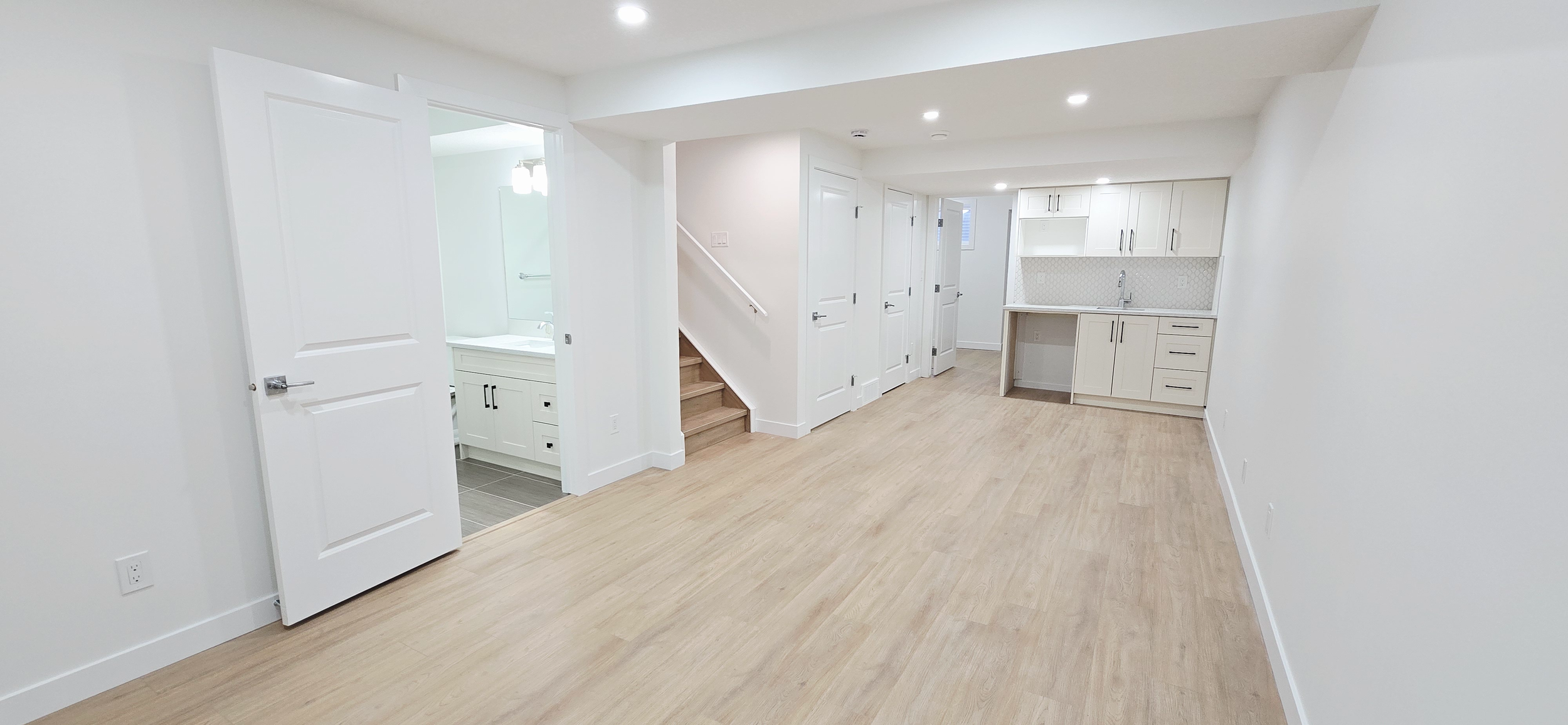 Bright modern basement suite with light oak vinyl plank flooring, white shaker kitchenette, recessed pot lights, and crisp white walls and doors.