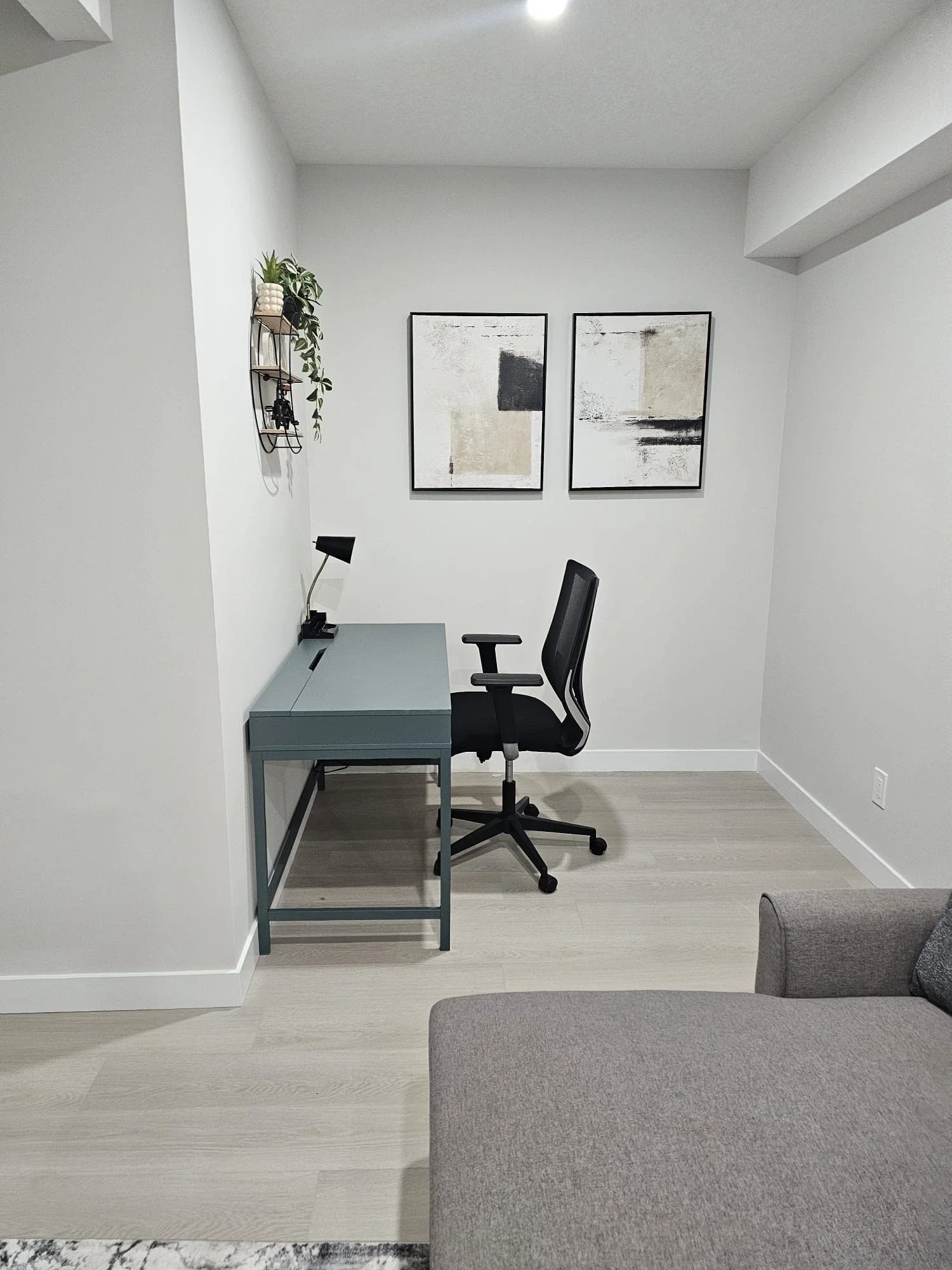Minimalist home office with a blue-grey desk, black mesh ergonomic chair, twin abstract art prints, and a hanging plant on light wood floors.