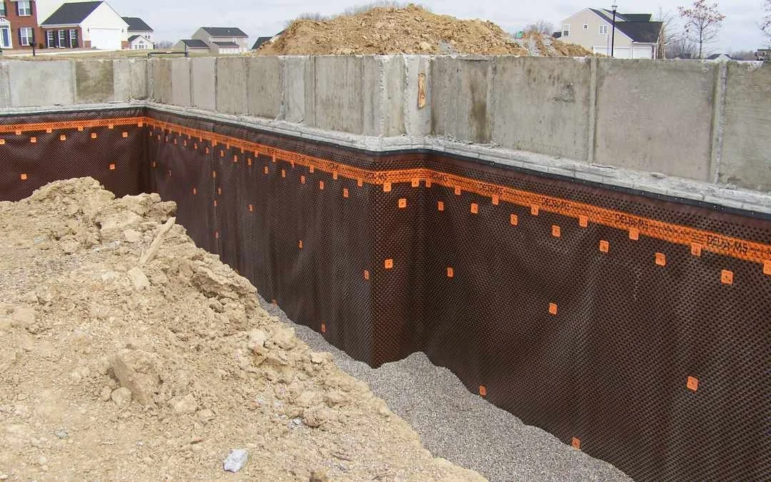 Description Generate Brown dimpled basement waterproofing membrane with orange trim on gray concrete walls above gravel drainage at a residential construction site.