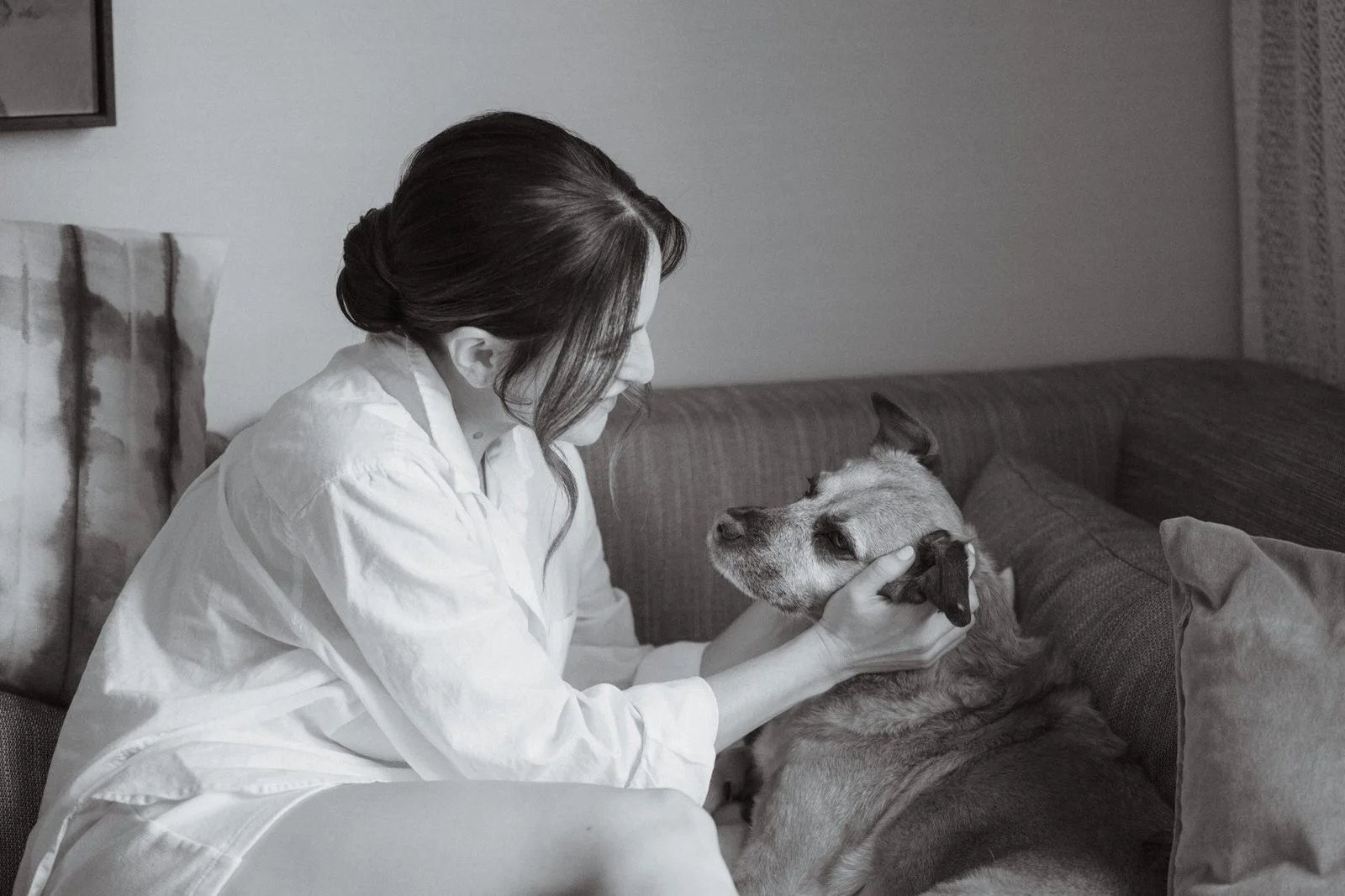A woman with dark hair in a bun gently holds a dog's face while sitting on a couch in a living room.