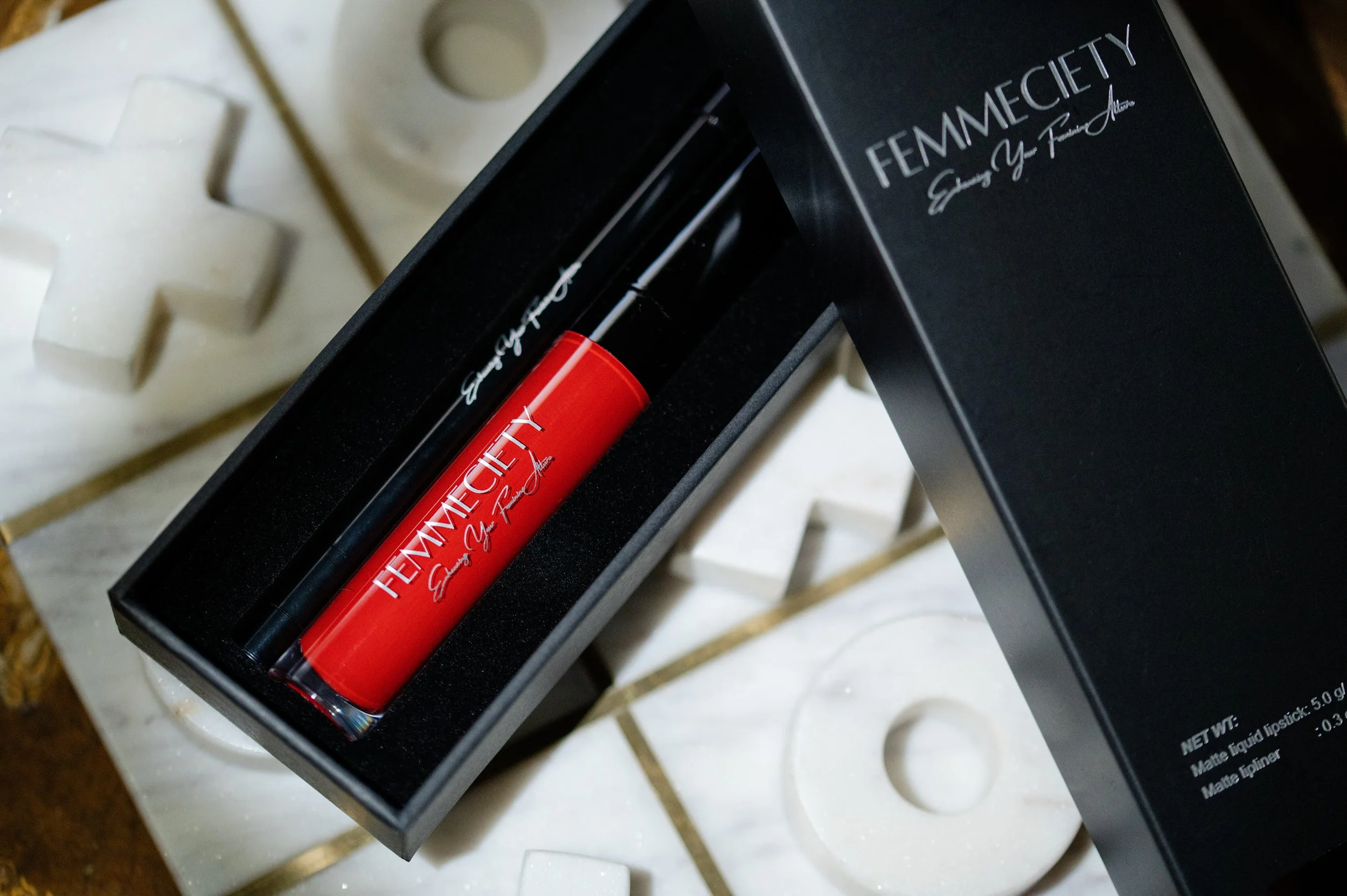 Open black box containing lipstick and lip gloss with 'FEMME CITY' branding, placed on a textured white surface with decorative white crosses.