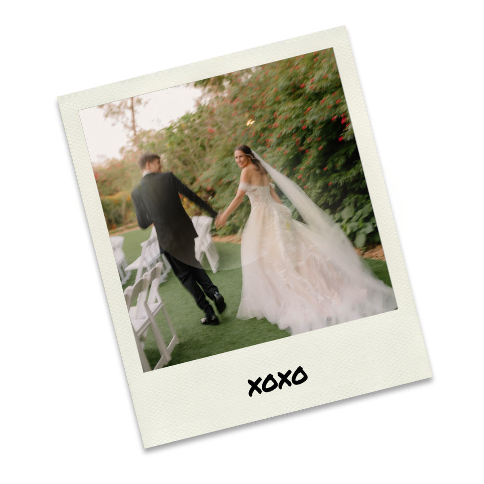 A bride and groom holding hands, walking on a grassy outdoor wedding venue surrounded by bushes and trees, with white chairs arranged nearby.