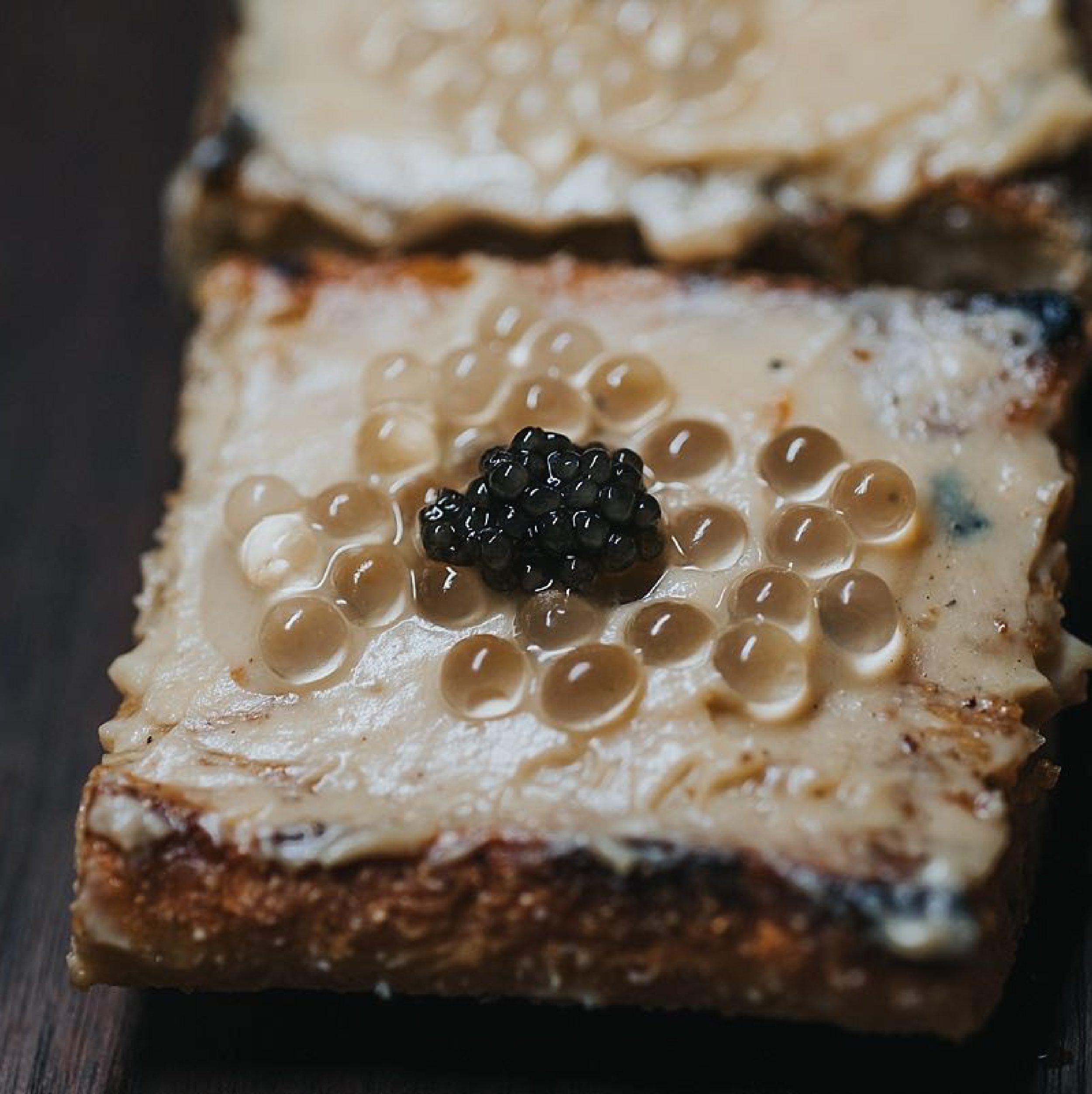 Close-up of a piece of bread with cream cheese and caviar topping, with black and beige caviar on top.