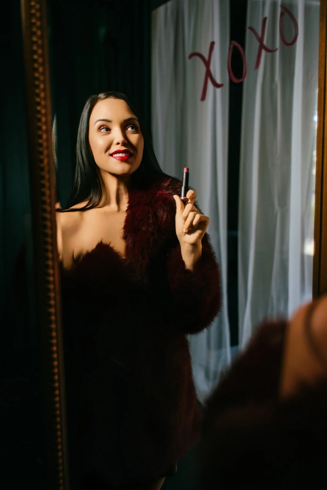 Woman with dark hair and red lipstick looking at herself in a mirror while holding a lipstick