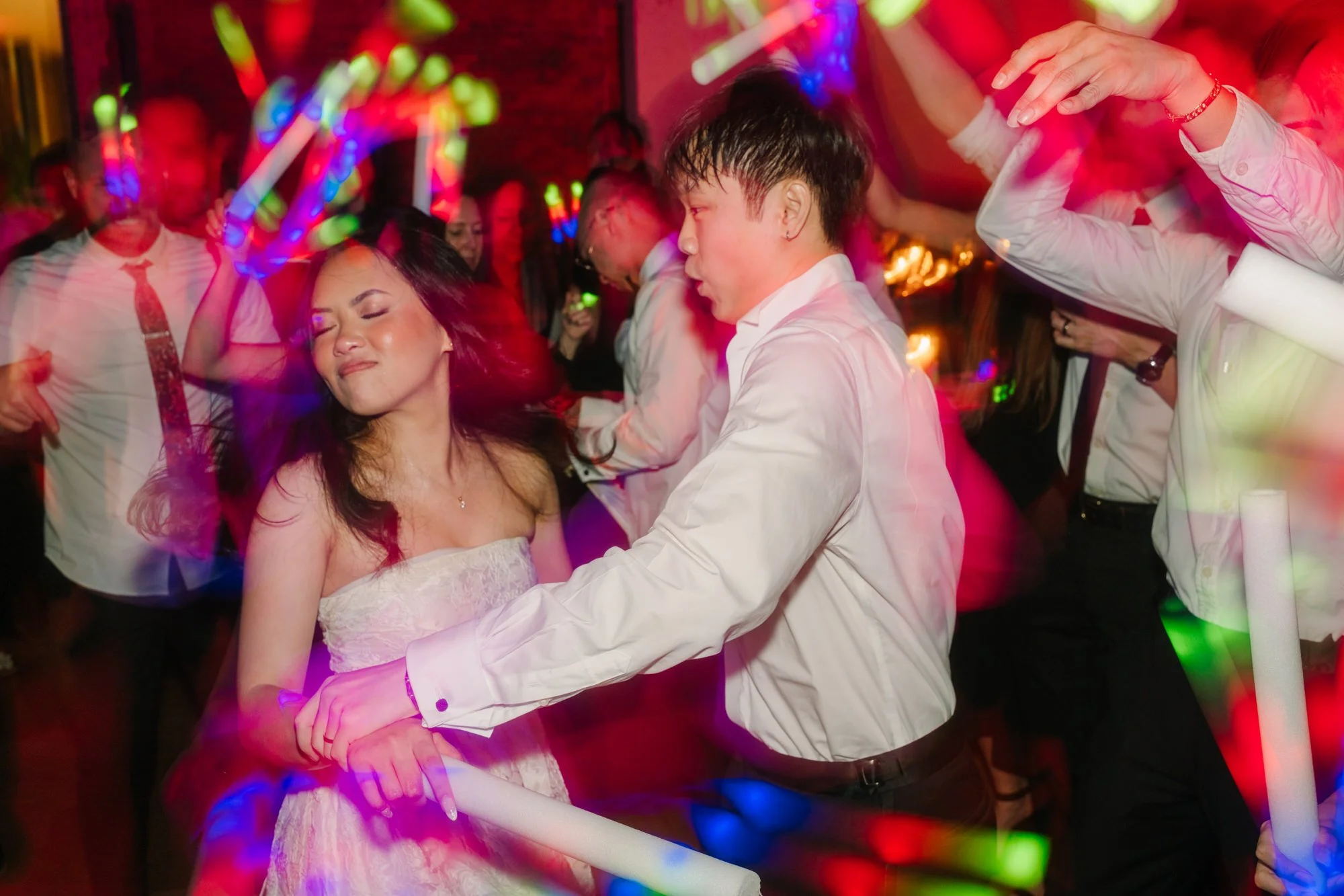 A bride and groom dancing closely at a wedding, with colorful lights and other people dancing in the background.