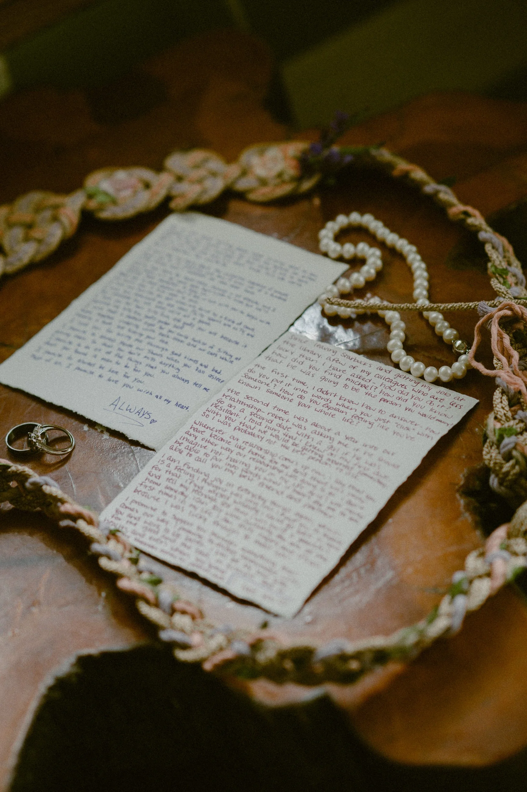 A handwritten wedding vow letter, pearl necklace, wedding rings, and floral braided fabric on a wooden table.