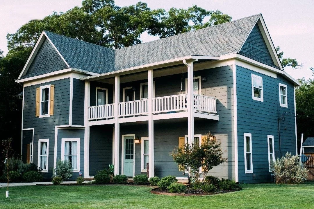 A two-story house with blue siding, white trim, and a gabled roof. It has a front porch with a balcony above, several windows with yellow shutters, and a green front door. The house is surrounded by a well-maintained lawn and landscaped bushes.