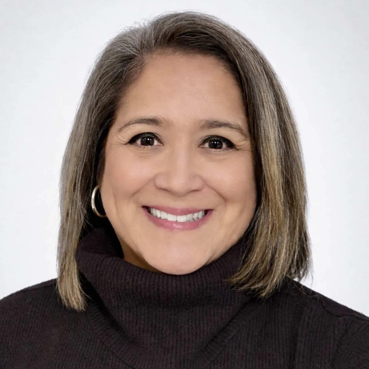 A woman with shoulder-length brown hair wearing a black turtleneck sweater and hoop earrings, smiling against a white background.