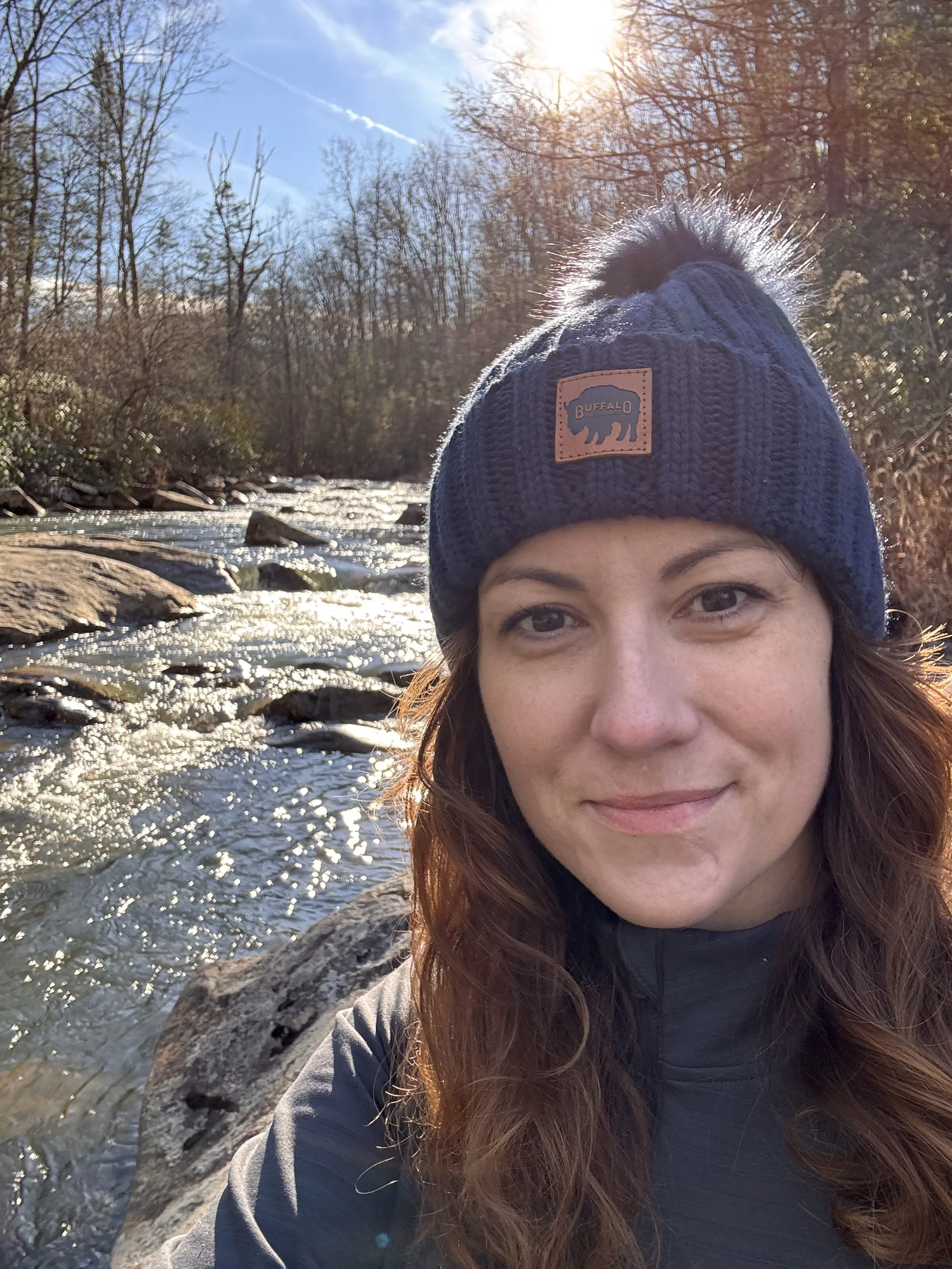 A woman outdoors near a flowing stream, wearing a blue knit beanie with a fur pom-pom and a jacket, with autumn trees and sunlight in the background.