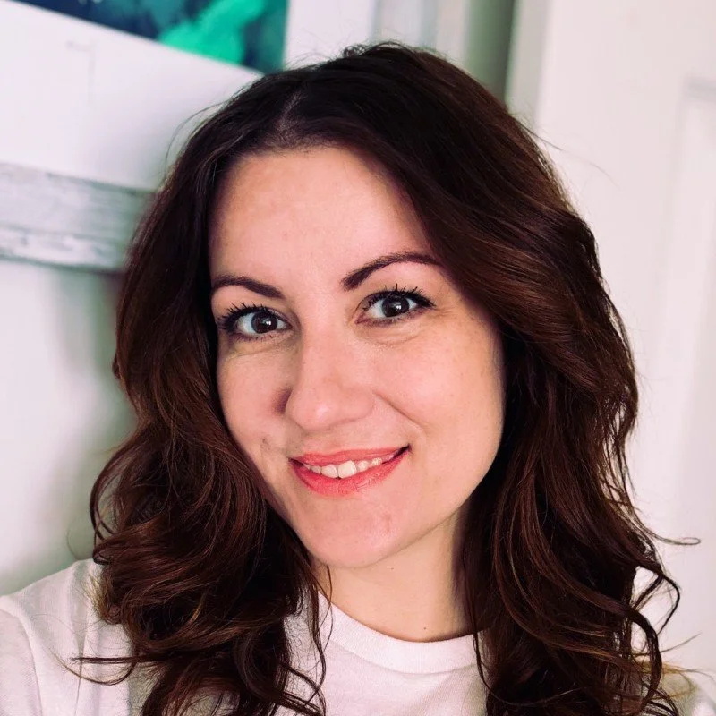 Close-up of a woman with dark brown wavy hair, smiling, wearing a white shirt, in an indoor setting with a light-colored background.