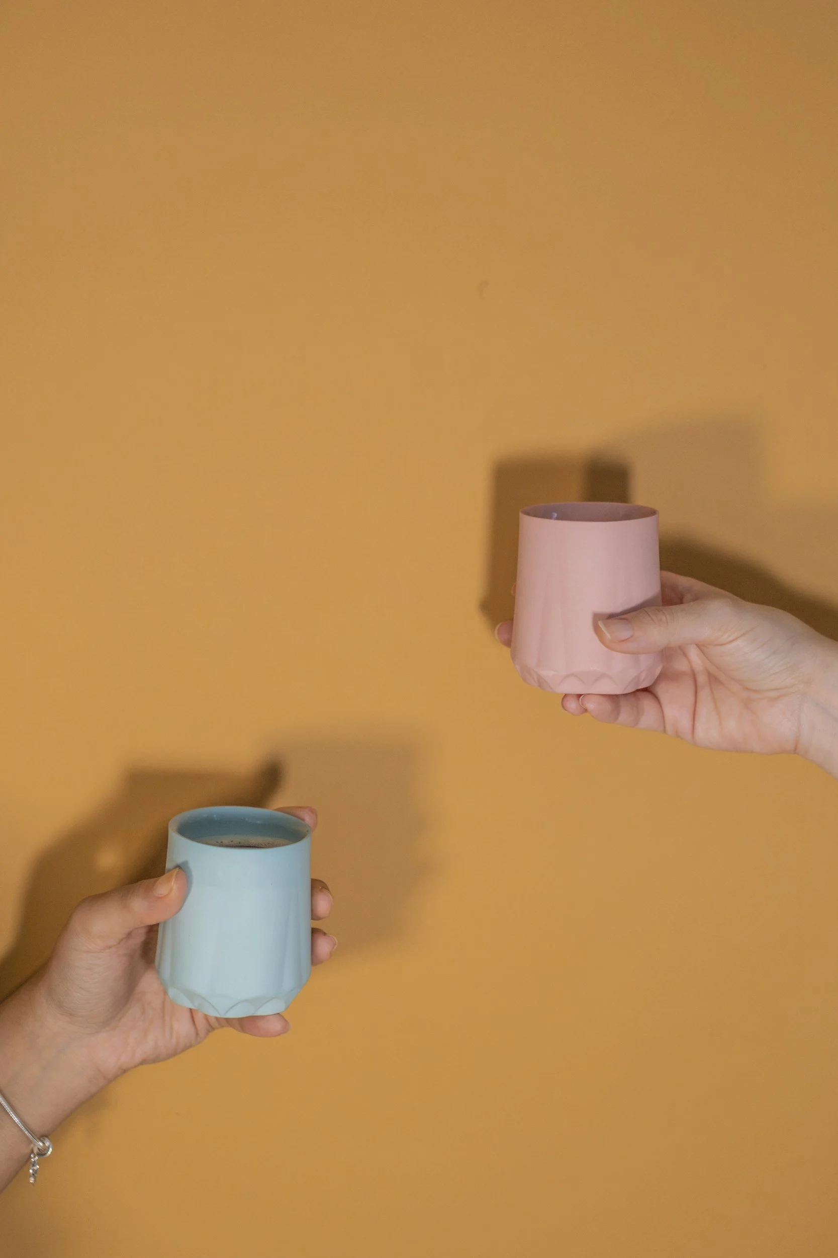 Two hands holding pastel-colored coffee cups against an orange background, with visible shadows of the cups on the wall.
