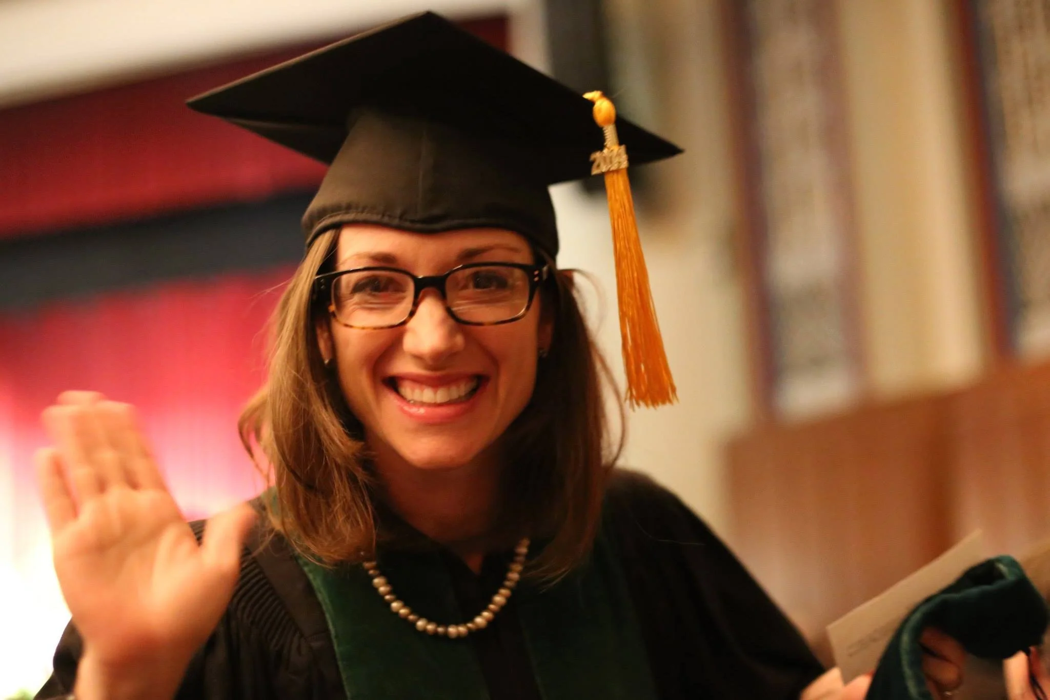 A woman in graduation cap and gown, smiling and waving at a graduation ceremony, wearing glasses and a pearl necklace.