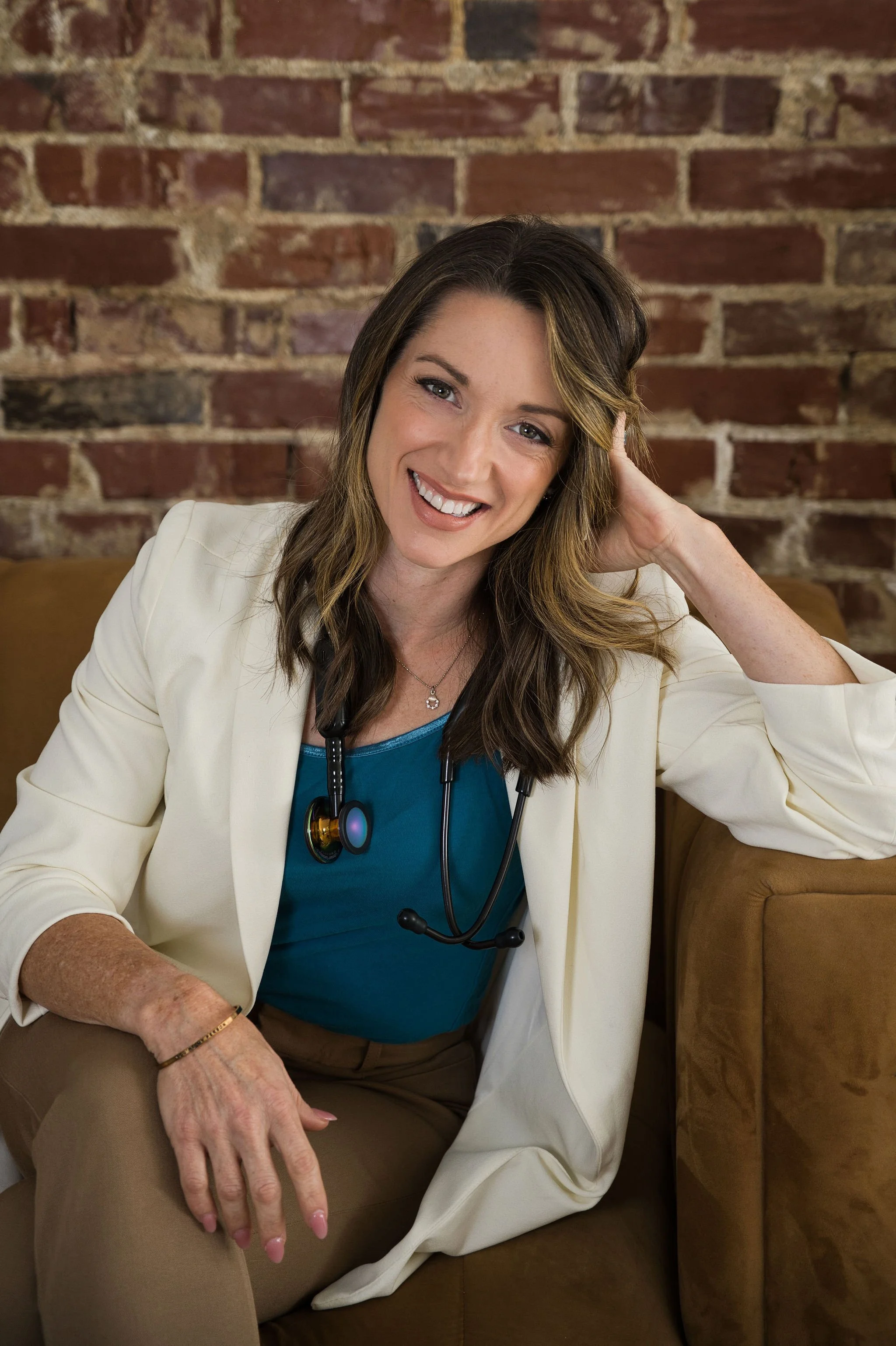 A woman with brown hair and a bright smile, wearing a white blazer, a teal top, and a stethoscope around her neck, seated on a brown sofa in front of a brick wall.